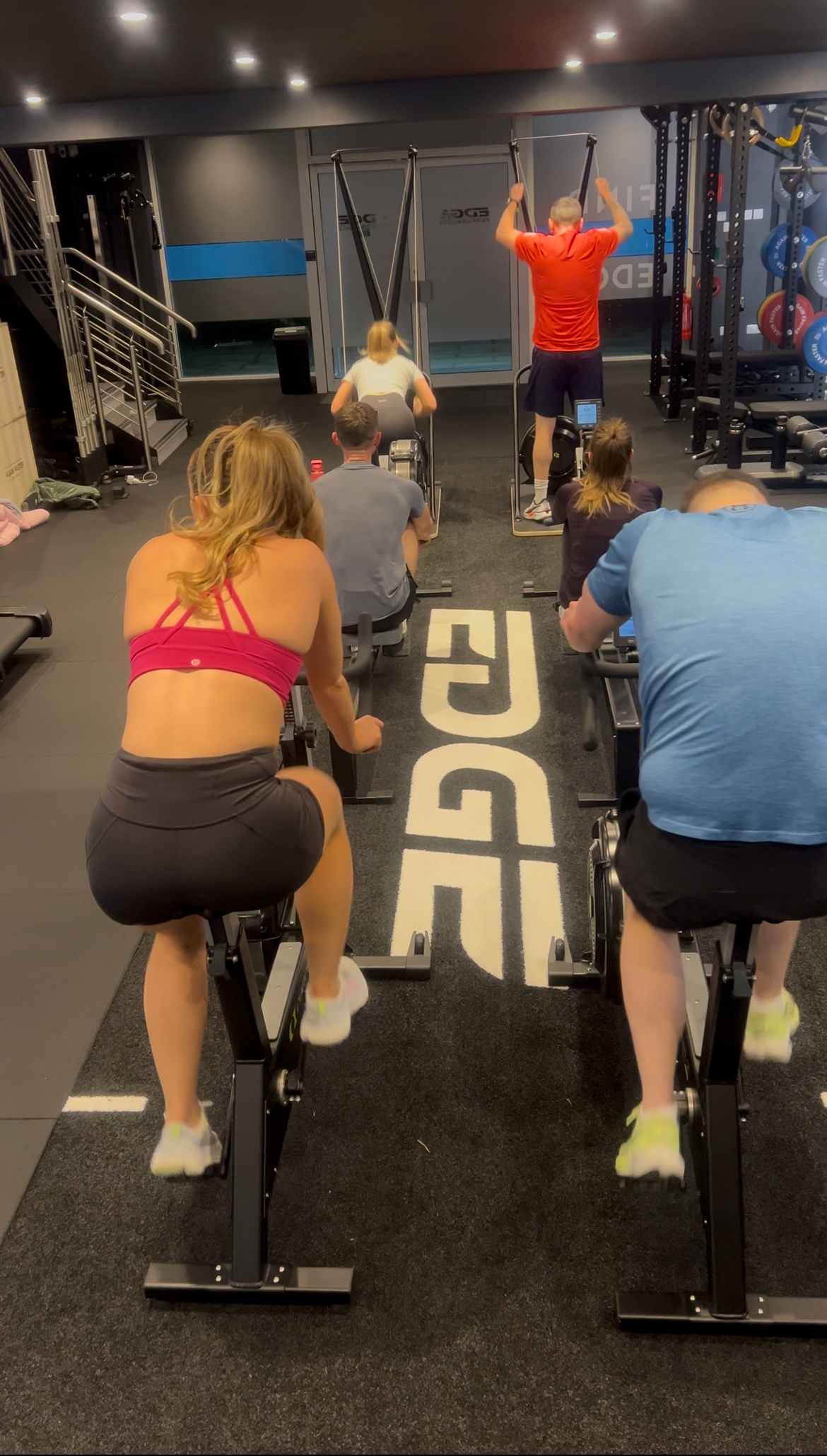 People working out on exercise bikes in a gym, with two individuals using resistance machines in the background and others seated on the bikes.