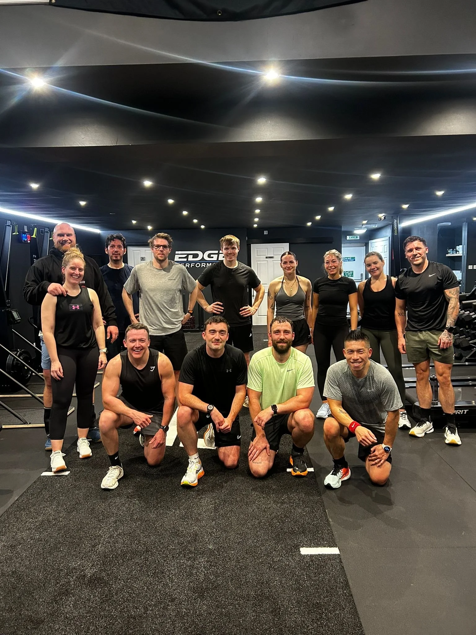 Group of 14 people in workout attire posing in a fitness gym with equipment and a black and gray color scheme.