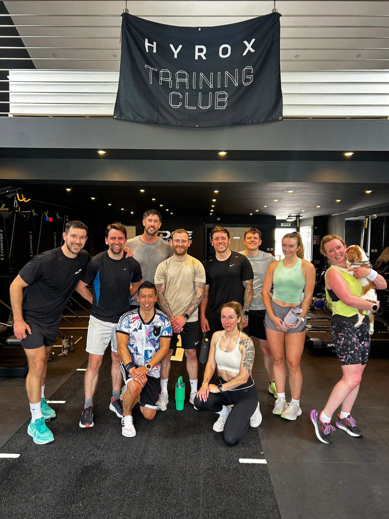 Group of nine people in workout clothes posing inside a gym with a banner reading 'HYROX TRAINING CLUB' hanging above.