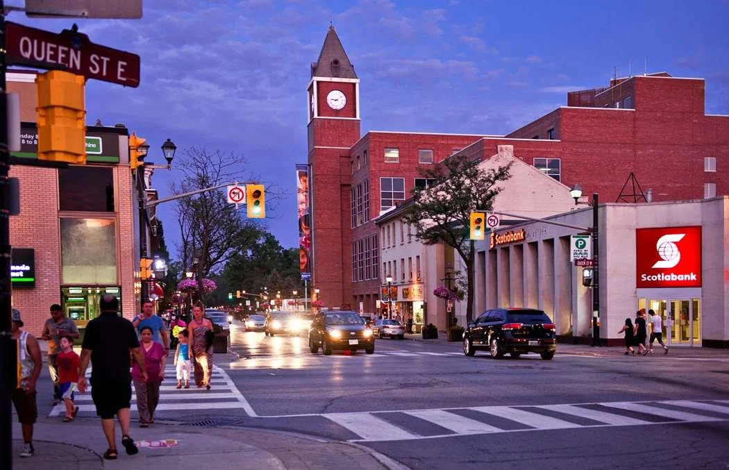 City street scene at dusk with pedestrians crossing a crosswalk, cars driving by, and a red brick building with a clock tower in the background. Street signs and a Scotiabank branch are visible.