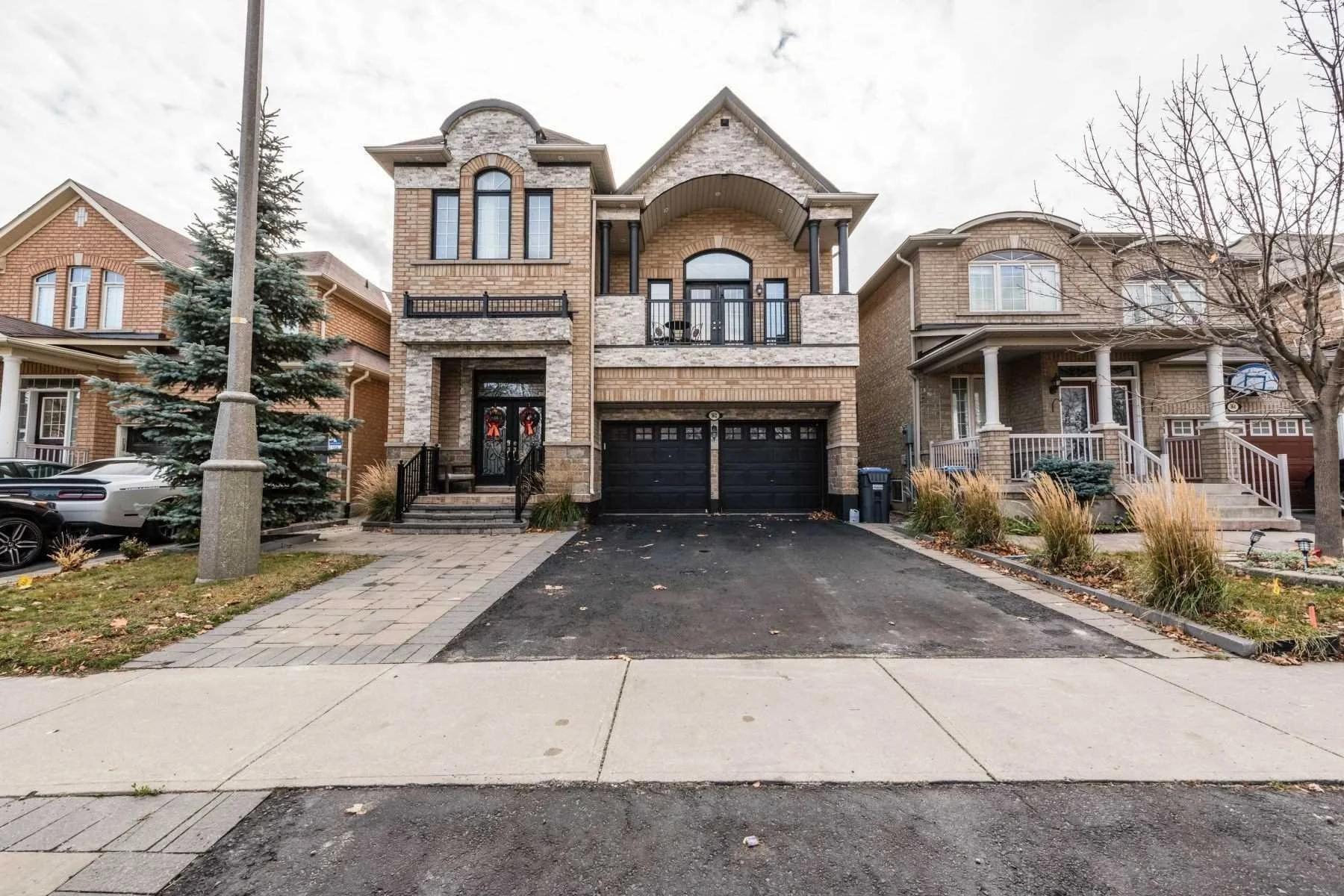 Front view of a multi-story brick house with a double garage, balcony, and steps leading to the front door, with neighboring houses and trees visible.