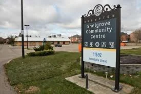 Sign for Snelgrove Community Centre on residential street, with grass and cloudy sky in background.