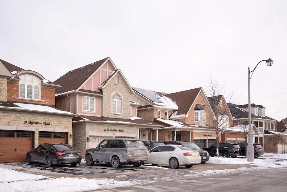 A row of suburban houses with garage doors, parked cars in front, and snow on the ground and roofs during winter.