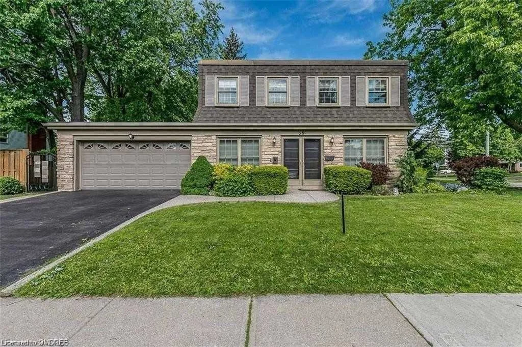 A two-story house with a stone and beige siding exterior, four upper windows, and a double front door. It has a two-car garage on the left with a dark driveway, and a well-manicured lawn with bushes and trees.