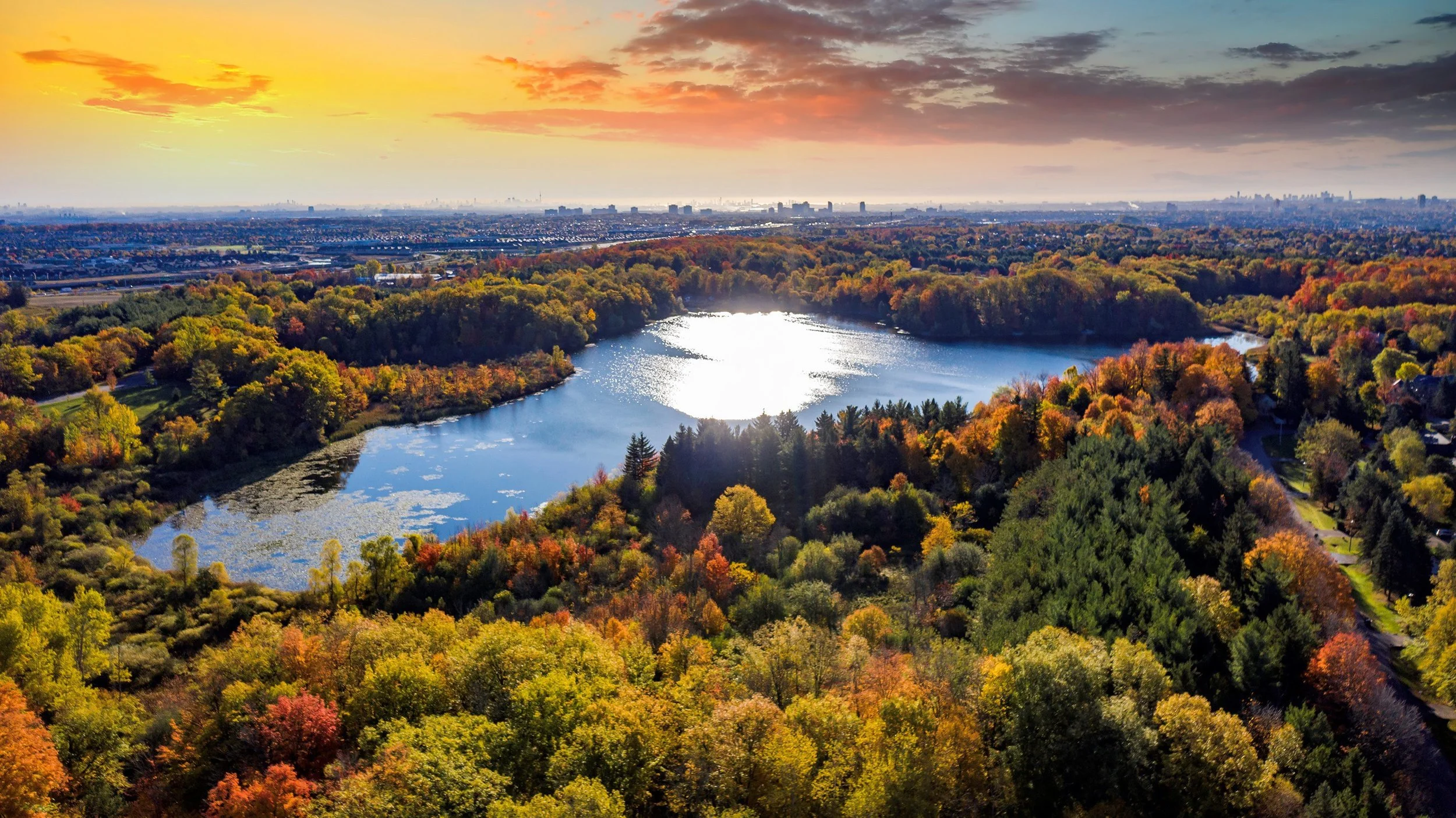 An aerial view of a forested area with a lake in the center, surrounded by colorful autumn trees, under a vibrant sunset sky with clouds.