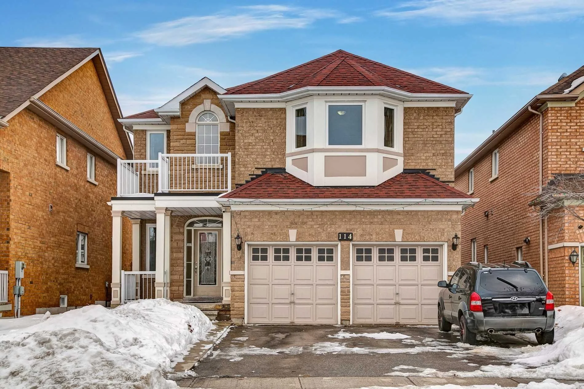 A two-story brick house with a red shingle roof, white garage doors, and a bay window. There is a black SUV parked in the driveway and snow on the ground.