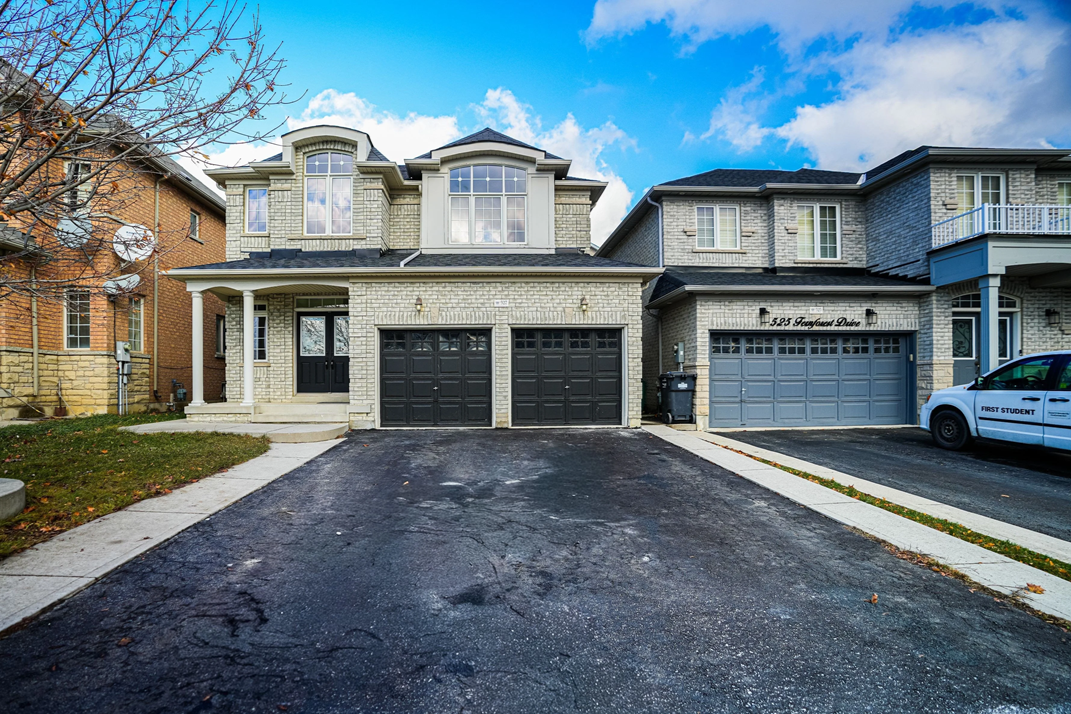 Two adjacent suburban houses with garages, driveway, and a sidewalk; the house on the left is beige brick with a small porch, the house on the right is gray brick with a balcony; blue sky with clouds overhead.