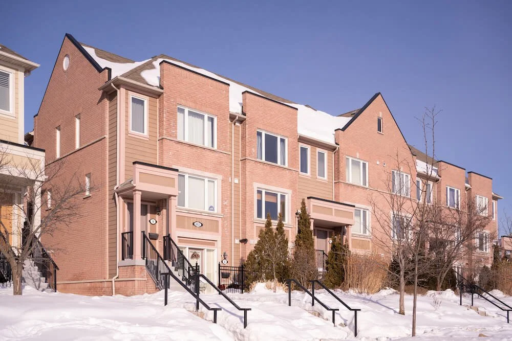A row of pink brick townhouses with snow-covered roofs and a snow-covered front yard, trees without leaves, black railings on the stairs, under a clear blue sky.
