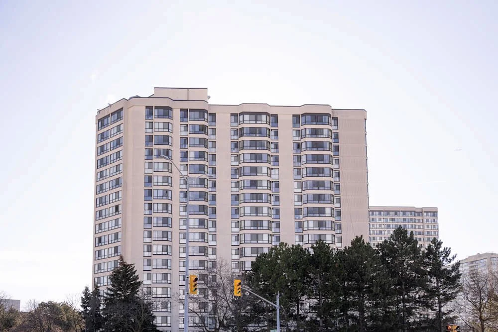 Tall residential apartment building with multiple floors and large windows, with trees and traffic lights at the street level in front.