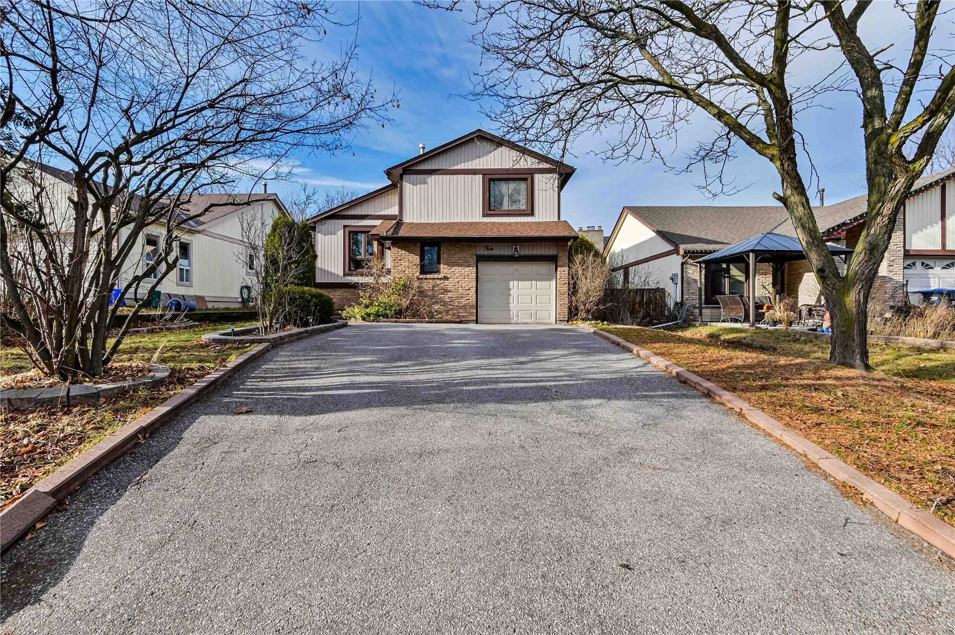 A residential house with a driveway, brick and siding exterior, and a garage door. There are leafless trees and neighboring houses on either side, with a patio on the right side.