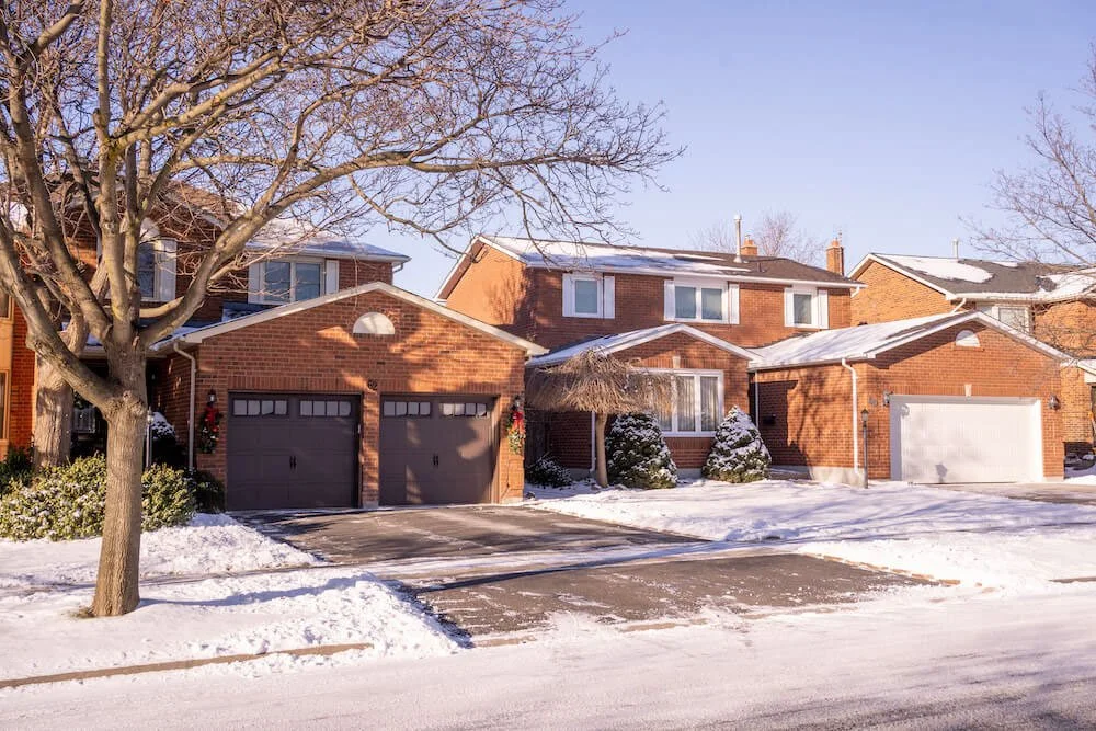 A suburban residential street with snow-covered ground and rooftops, brick houses, and trees with no leaves under a clear blue sky.