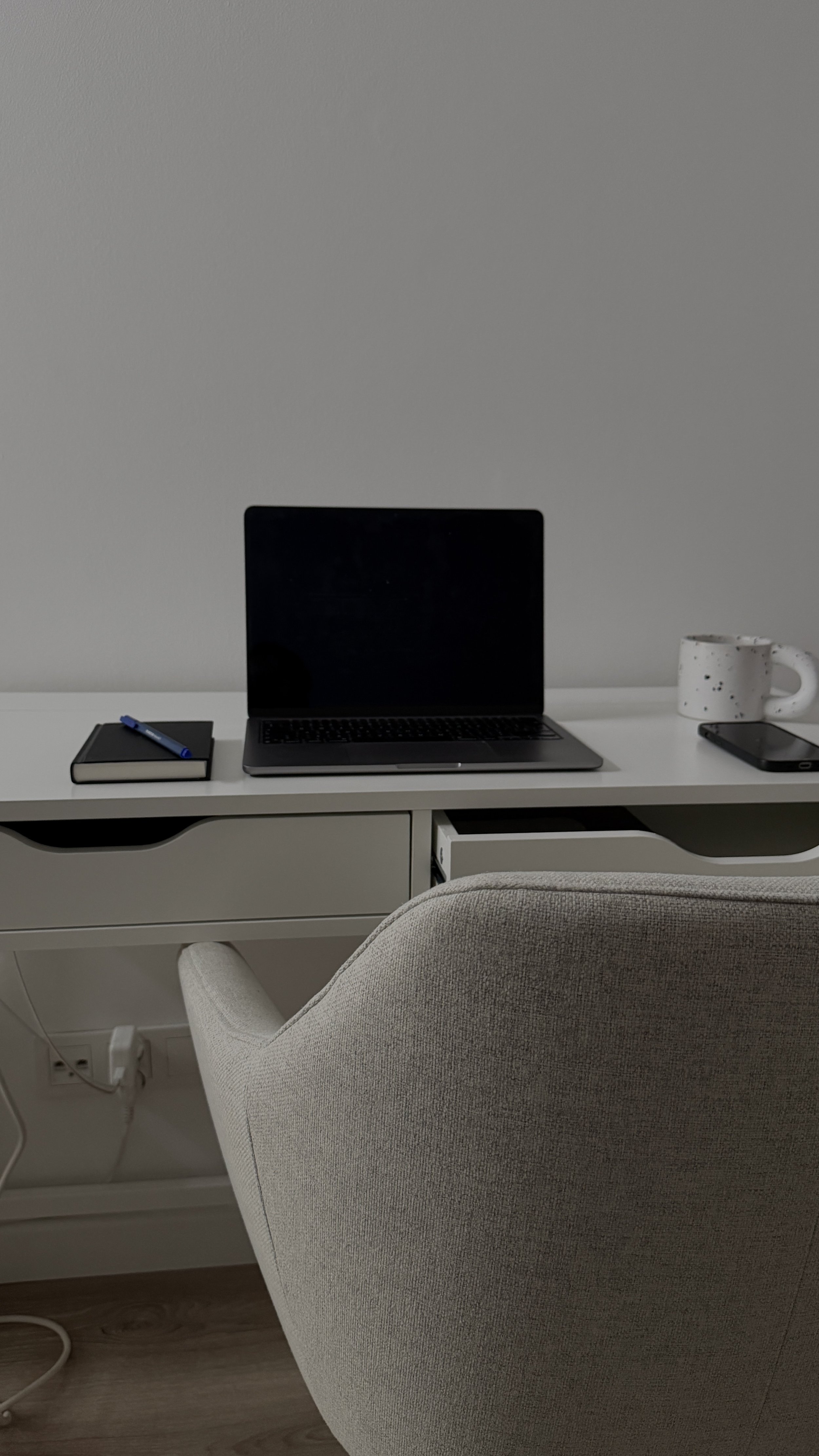 A white desk with a black laptop, a closed notebook with a pen on top, a ceramic mug, and a smartphone. A gray upholstered chair is in front of the desk, and a white wall is in the background.