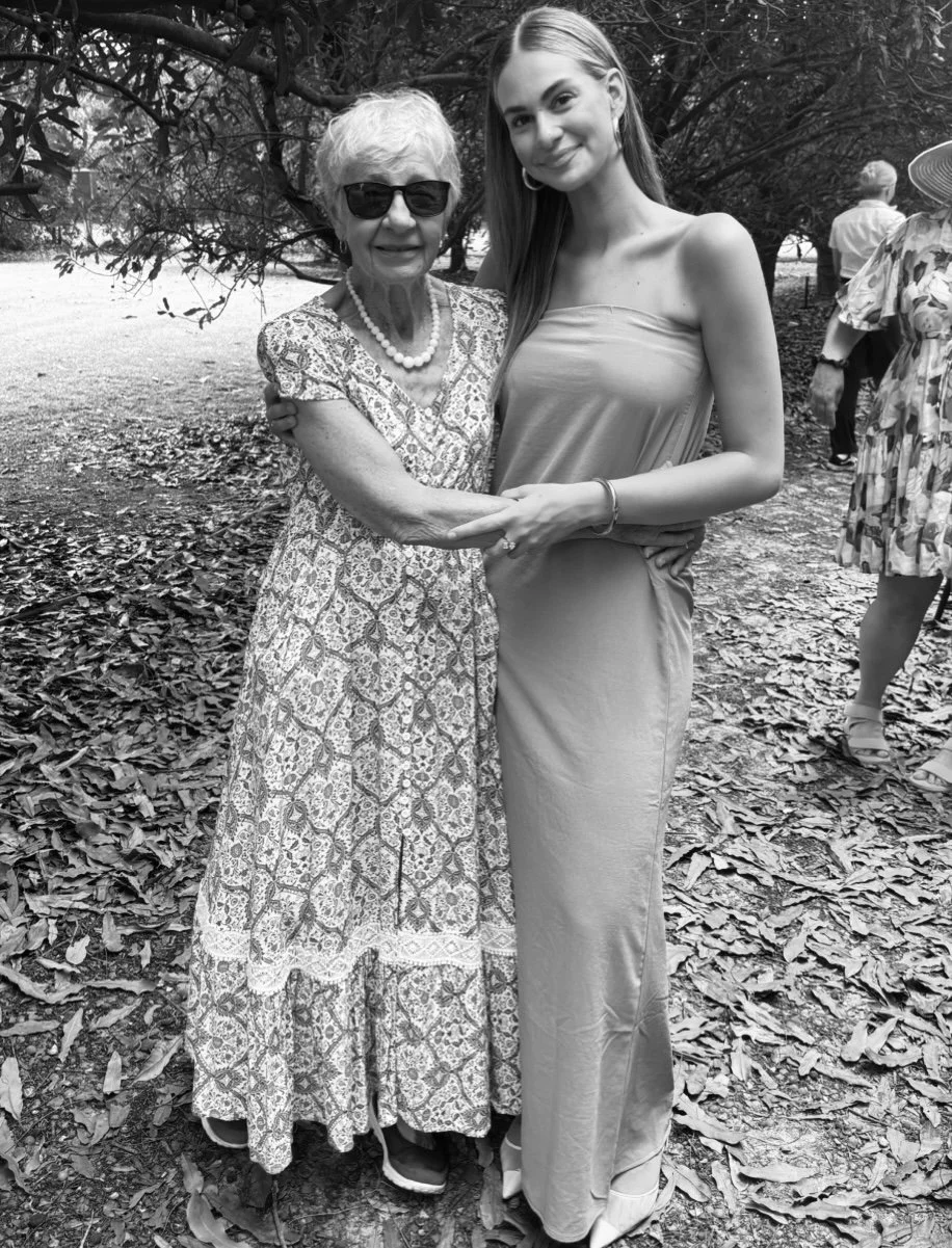 An elderly woman and a young woman standing together outdoors, smiling and holding hands. The elderly woman is wearing sunglasses, a patterned dress, and a pearl necklace. The young woman is in a strapless dress. There are other people in the background under a tree covered with leaves.
