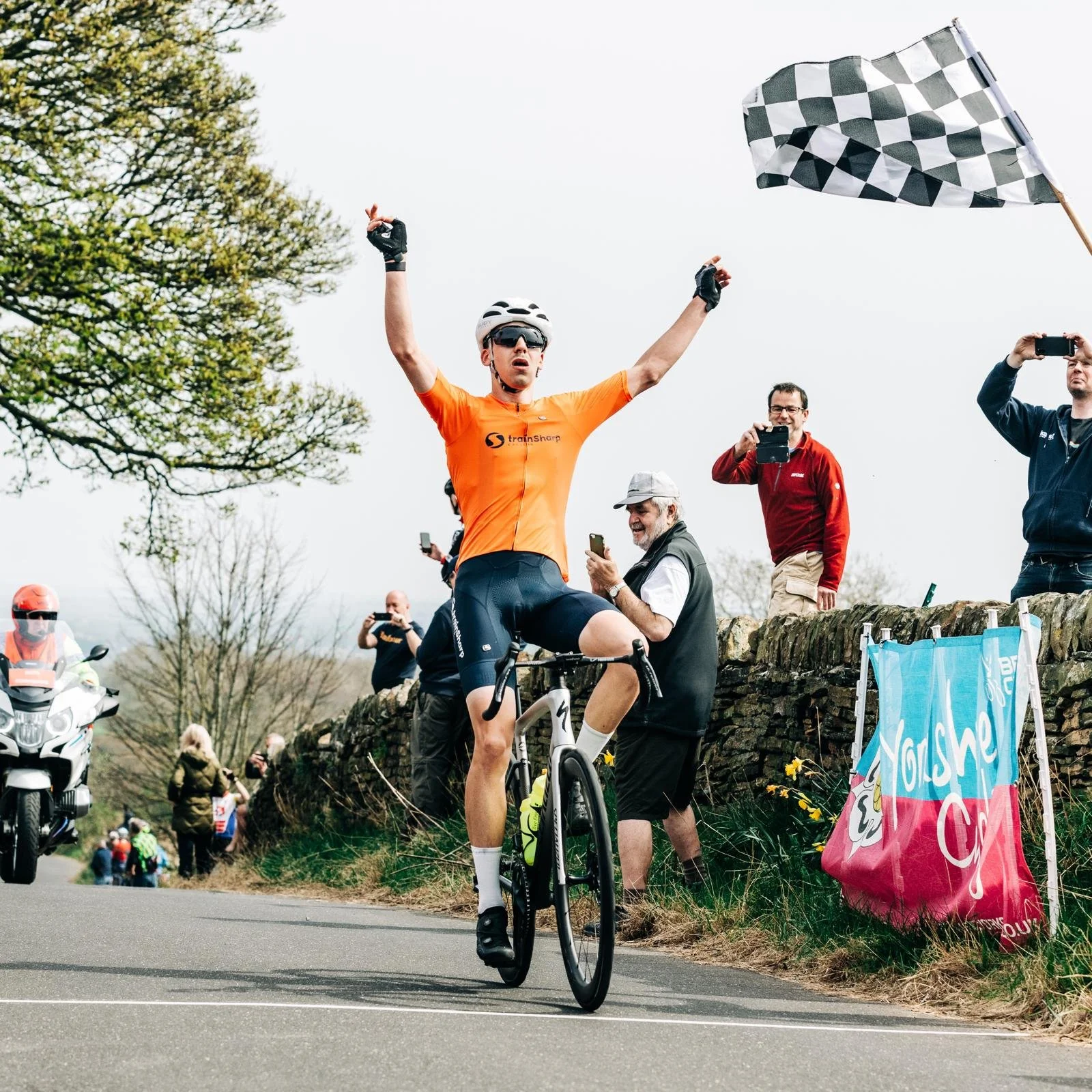 A cyclist in an orange jersey with arms raised celebrates as he crosses the finish line, surrounded by spectators and photographers on the roadside, with a checkered flag in the air.