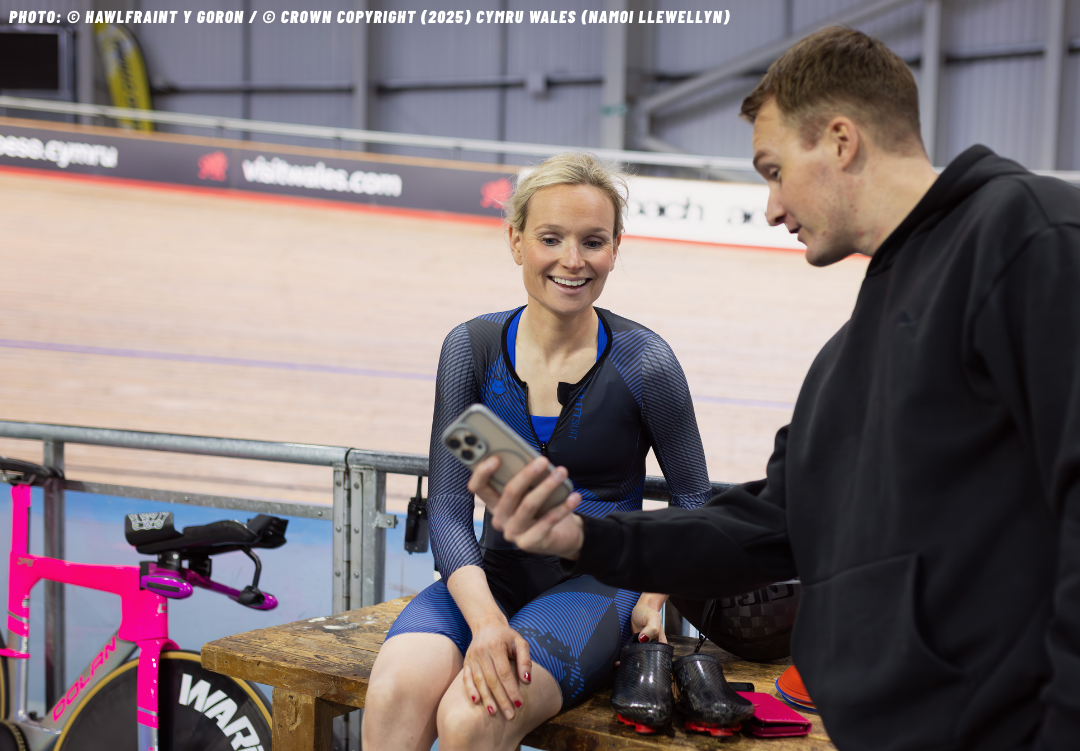 A woman in athletic gear sitting on a wooden bench at an indoor velodrome, smiling as a man shows her something on his phone. A pink bike is beside the bench, and cycling shoes and a helmet are on the bench.