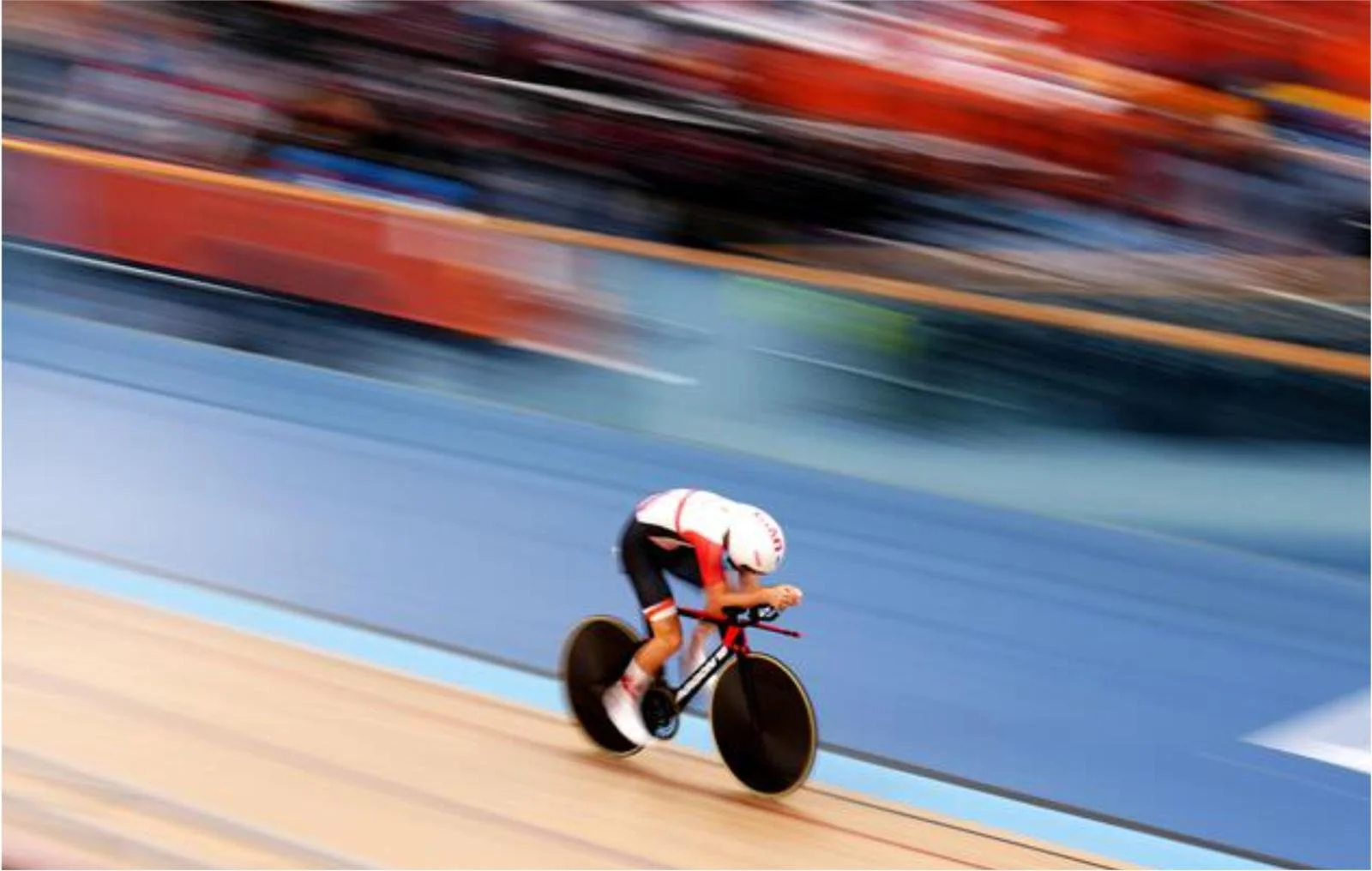 A cyclist wearing a white helmet and red and white uniform riding a black and red racing bike on an indoor velodrome track with colorful blurred background.