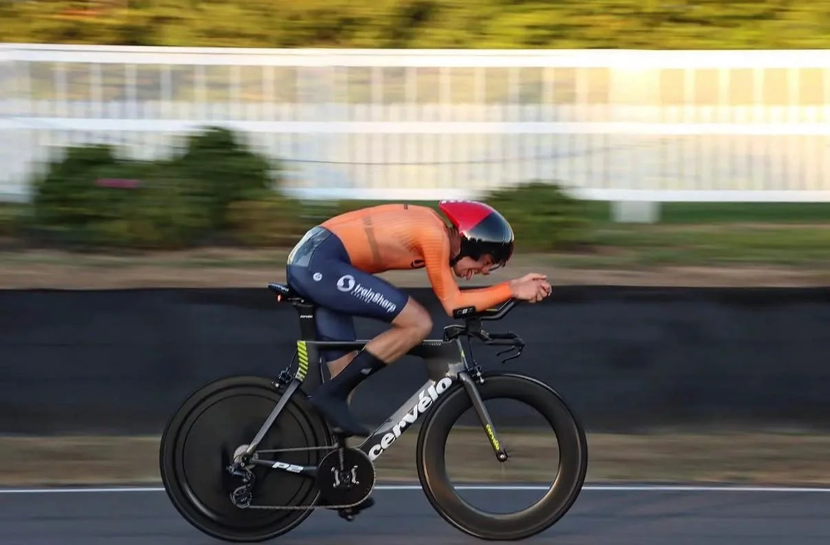 A cyclist wearing an orange jersey and black shorts rides a black Cervélo bike with aero wheels, while wearing a black and red helmet, on a paved road with blurred green bushes and a white fence in the background.