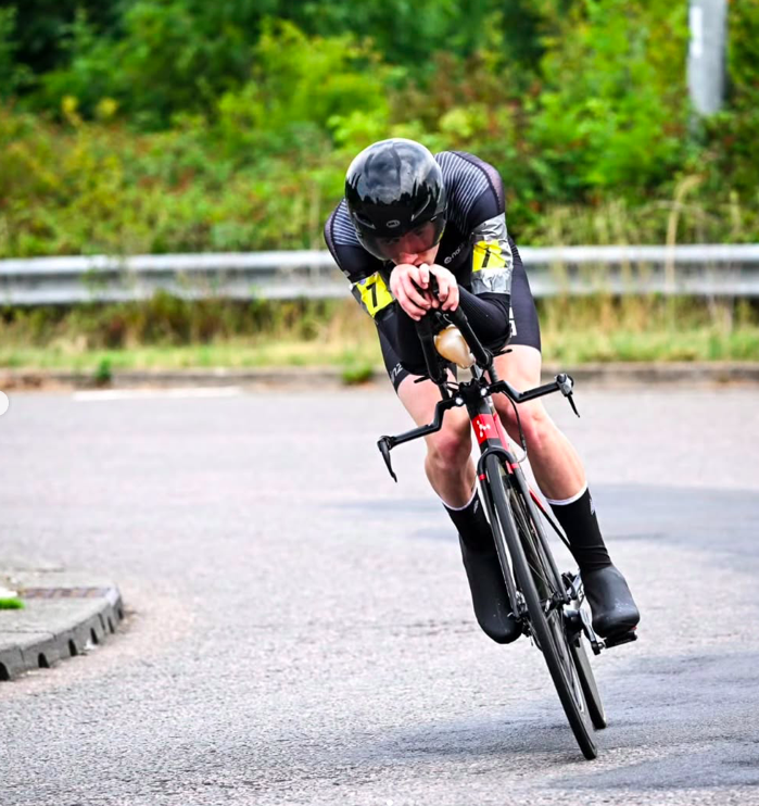 A cyclist, wearing a black helmet and black cycling gear, leans into a turn on a paved road during a race.