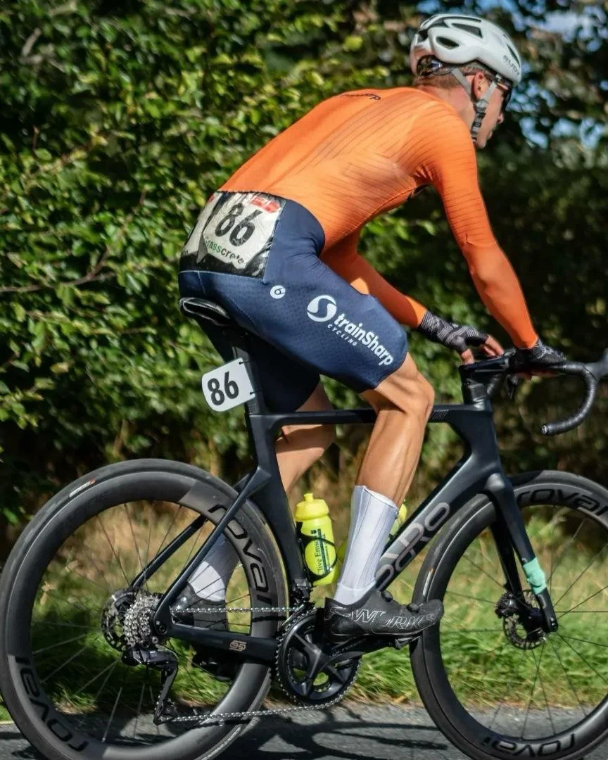Cyclist wearing orange jersey, black shorts, helmet, and sunglasses riding a black Specialized S-Works bicycle with a rider number 86 attached, on a road with green trees in the background.