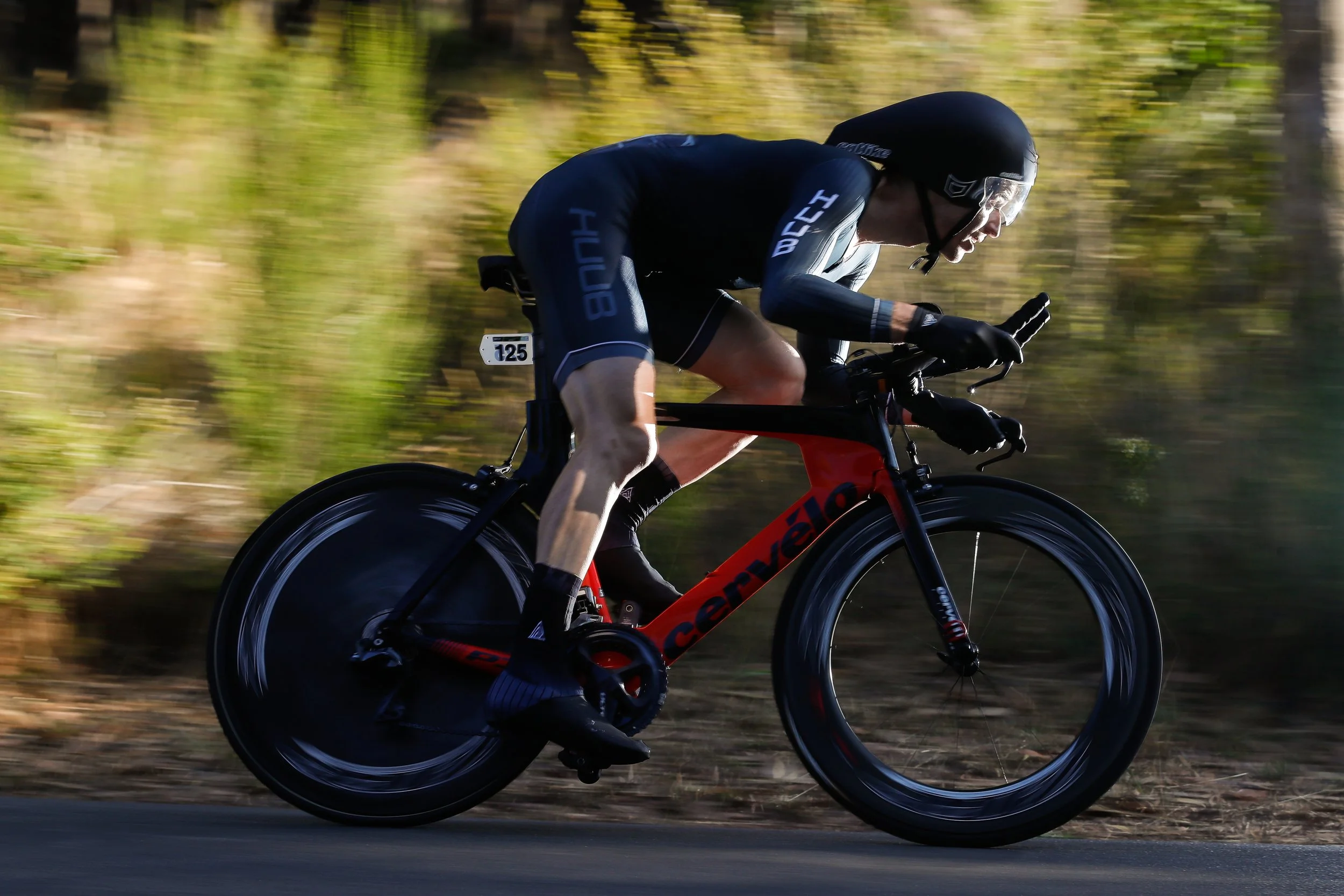 A cyclist wearing a black helmet and black cycling suit riding a red Cannondale race bike on a road with blurred greenery in the background.