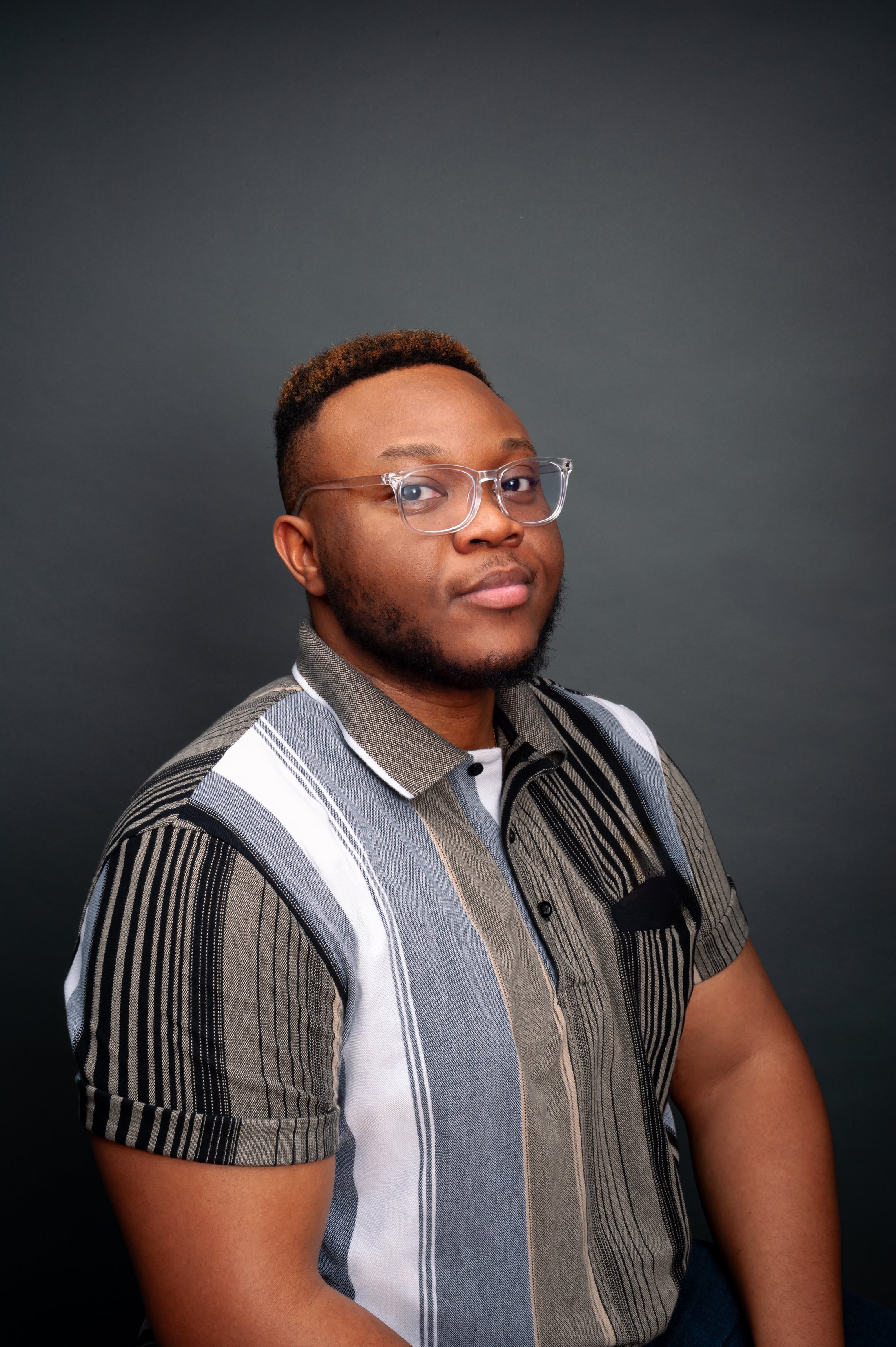 A young man with glasses, short hair, and a beard sitting against a dark gray background, wearing a short-sleeved, striped button-up shirt.