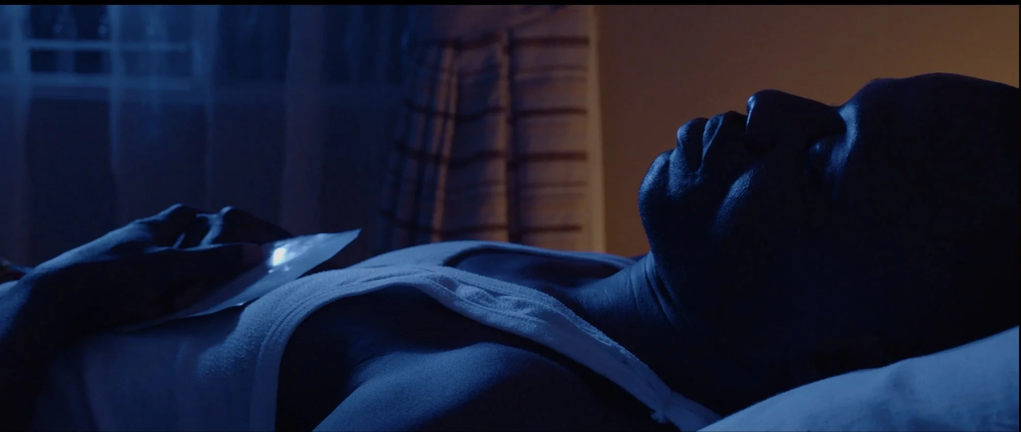 A person lying on their side in bed at night, holding a smartphone, with a dimly lit background and striped curtains.