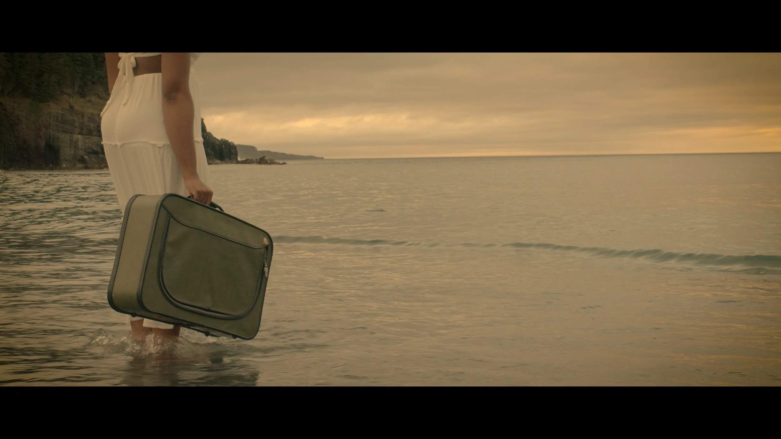 A person standing in shallow water at the beach, holding a green suitcase, with cliffs and a cloudy sky in the background.