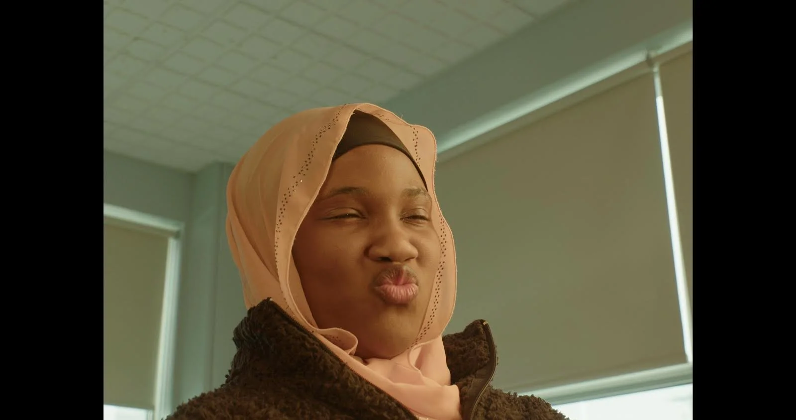 A woman making a duck face expression, wearing a peach-colored hijab and a brown fuzzy jacket, indoors in front of a window with closed blinds.
