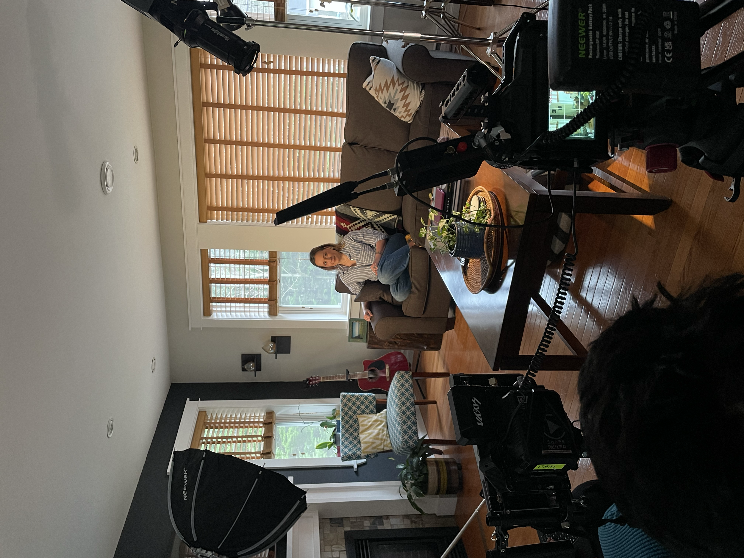 A woman sitting on a beige couch during a video recording or interview in a living room, with filming equipment including a camera, lights, and a boom microphone visible in the foreground.