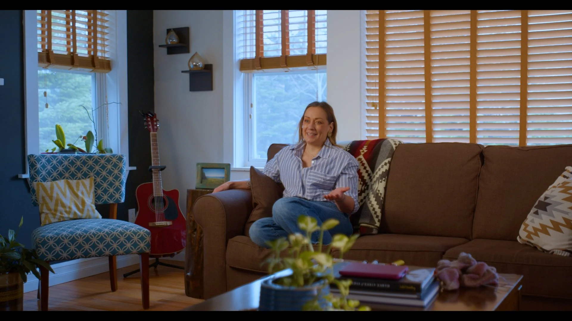 A woman sitting on a brown sofa, talking and smiling in a living room with large windows, wooden blinds, a guitar, a patterned chair, and plants.