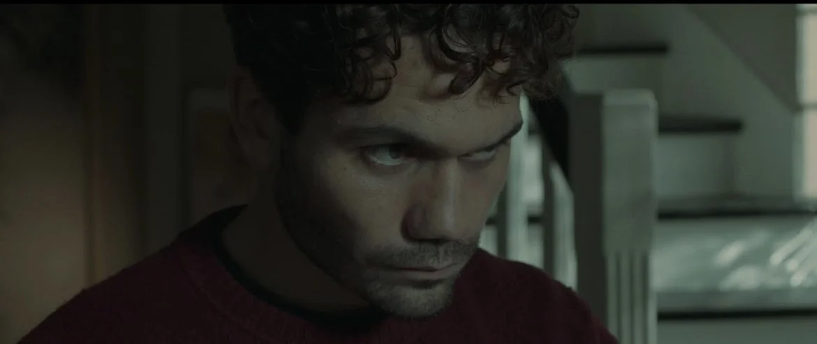Close-up of a young man with curly hair and intense expression, looking downward inside a dimly lit room.