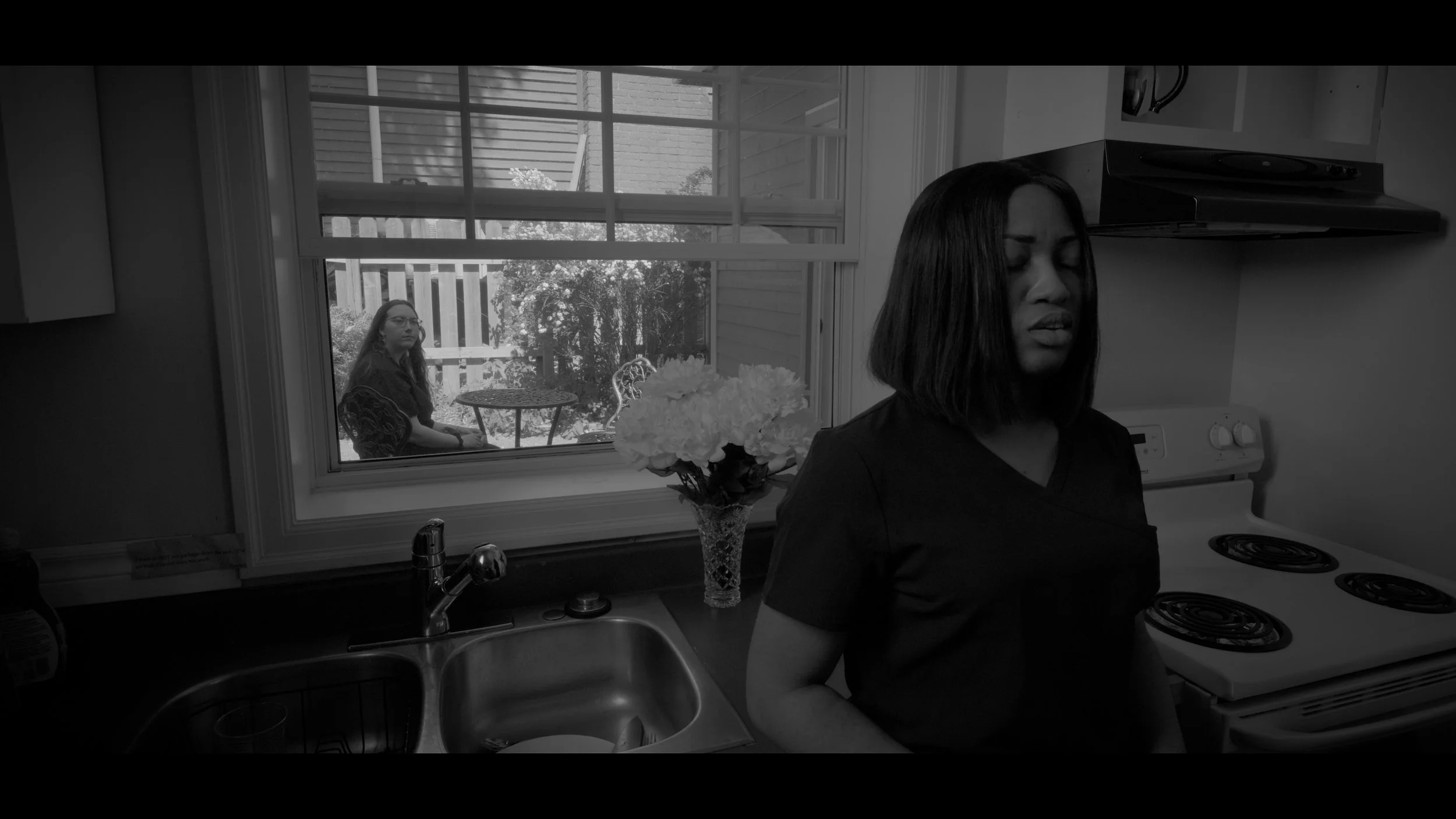 A woman standing in a kitchen with her eyes closed, and a young woman sitting outside on a patio, visible through a window.