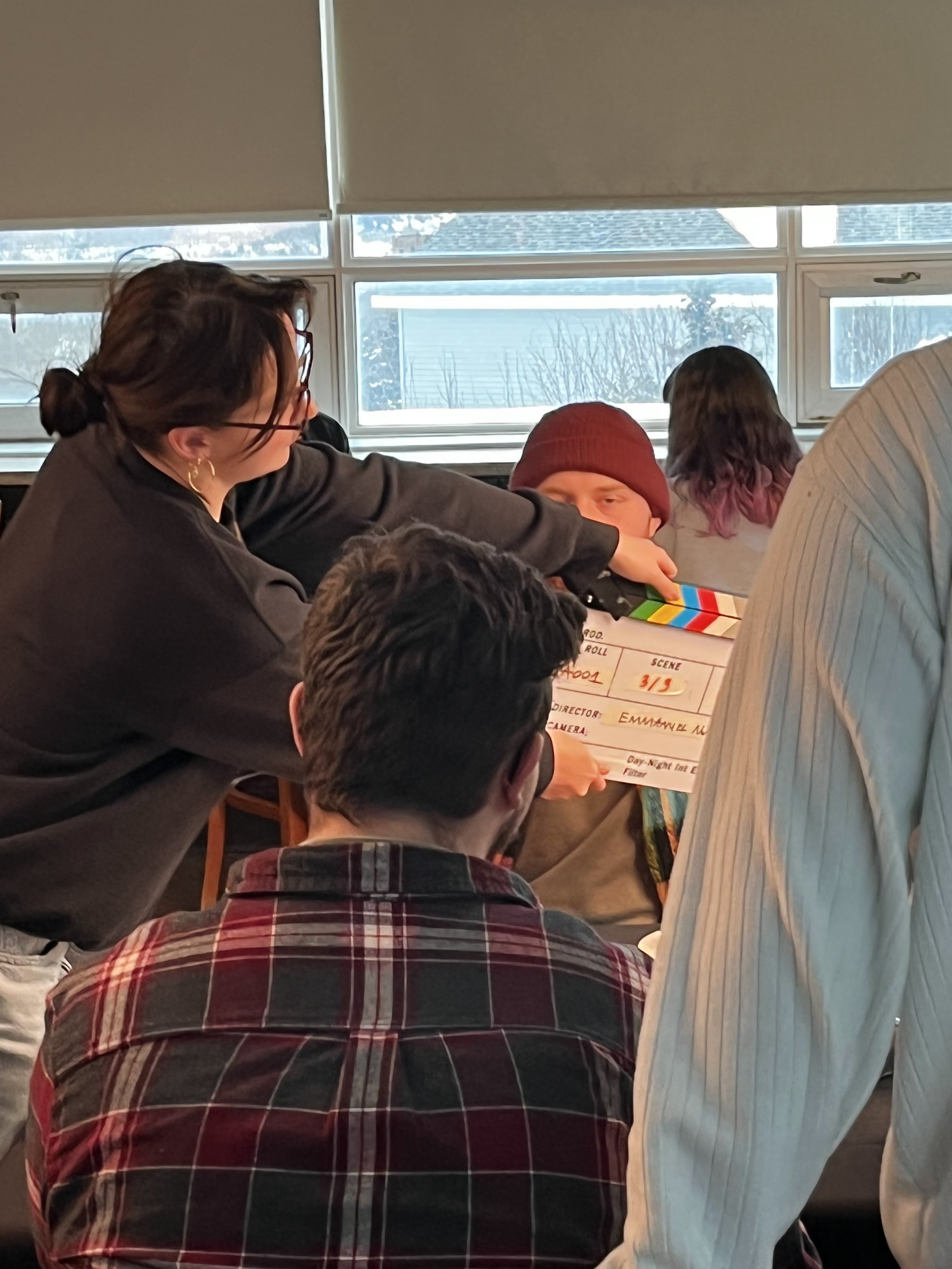 A woman in glasses and a dark top is holding a clapperboard for a film scene, and a man with a maroon beanie is sitting nearby. There are other people and a window in the background.