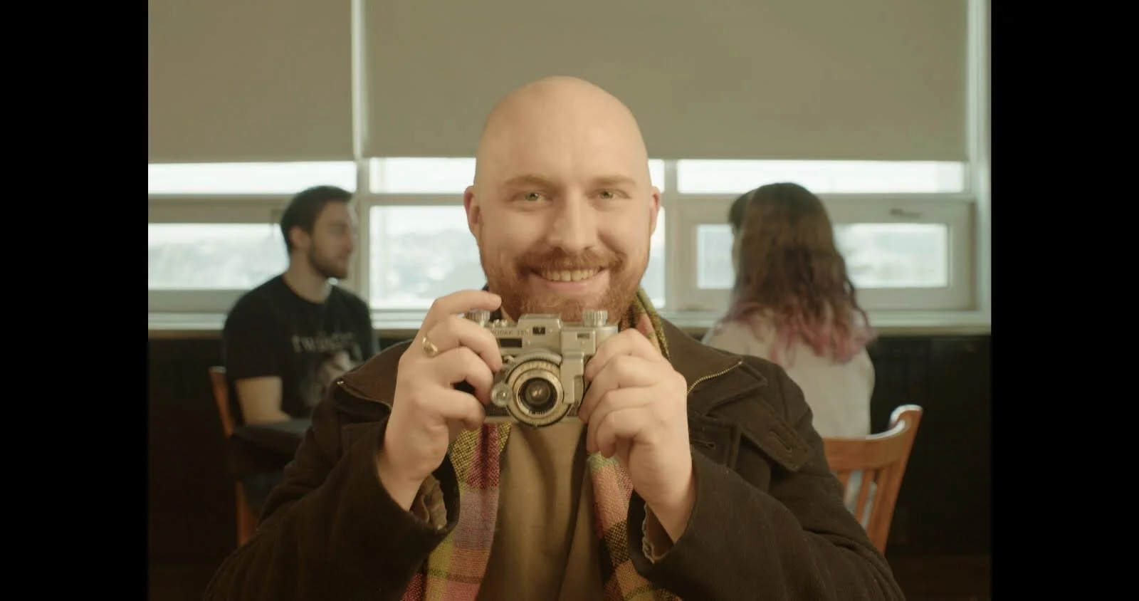 A smiling man with a beard and shaved head holding a vintage camera indoors, with two people sitting at a table and windows in the background.