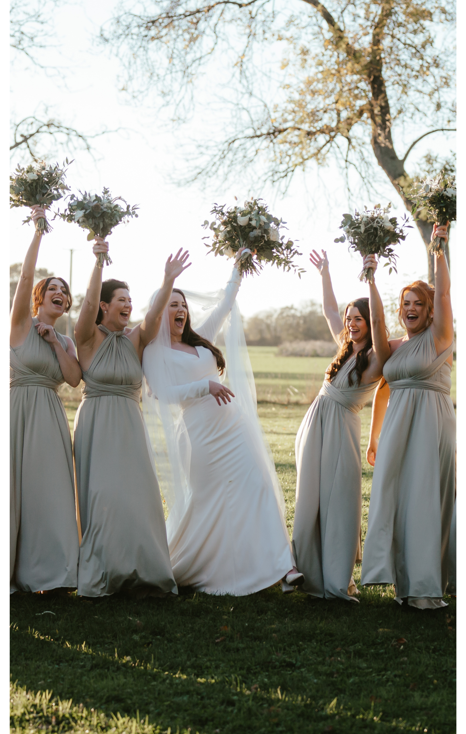 Bride and five bridesmaids celebrating outdoors, holding bouquets, smiling, with trees in the background.