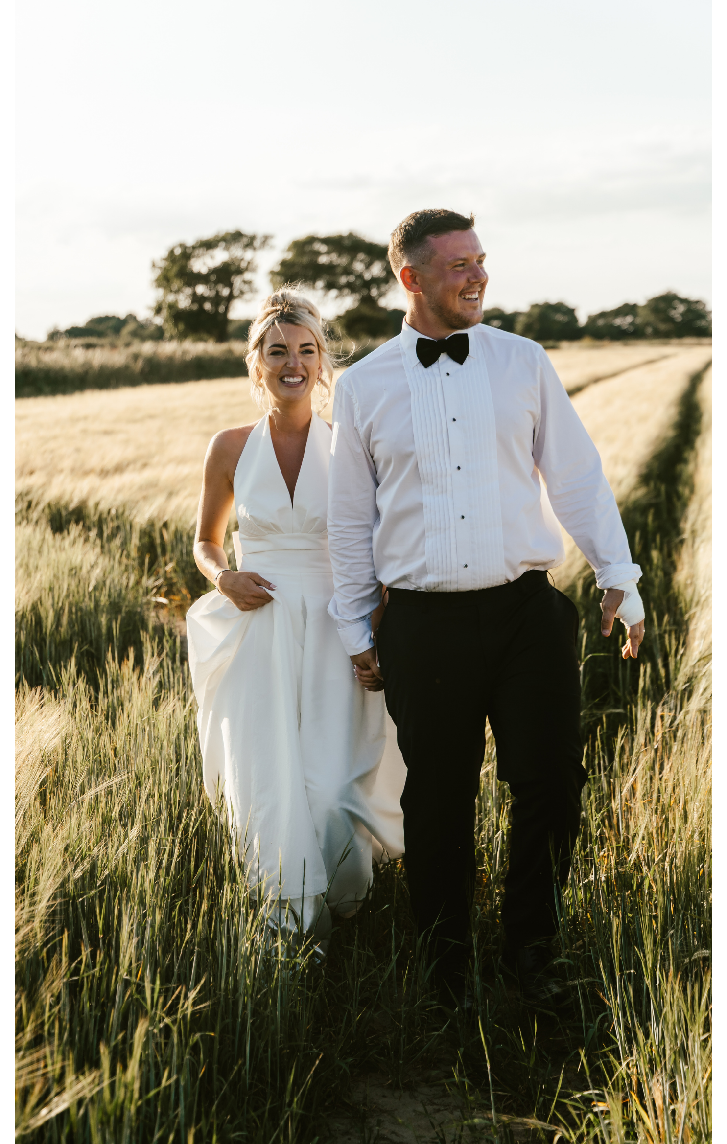 A newlywed couple walking hand-in-hand through a wheat field, smiling and looking joyful, with trees and a cloudy sky in the background.