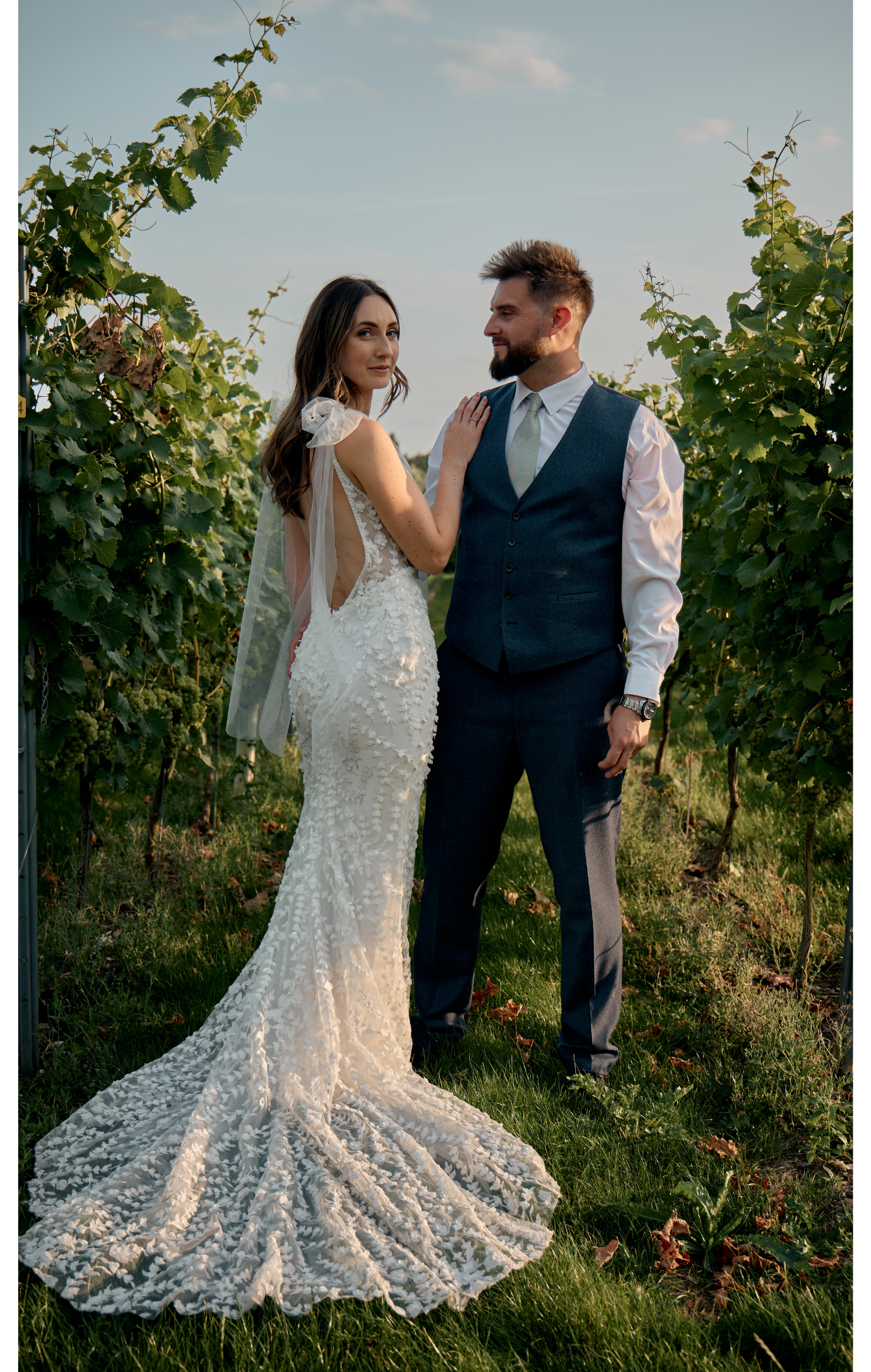 A bride and groom in wedding attire standing close together outdoors in a vineyard, with lush green grapevines around them, under a partly cloudy sky.