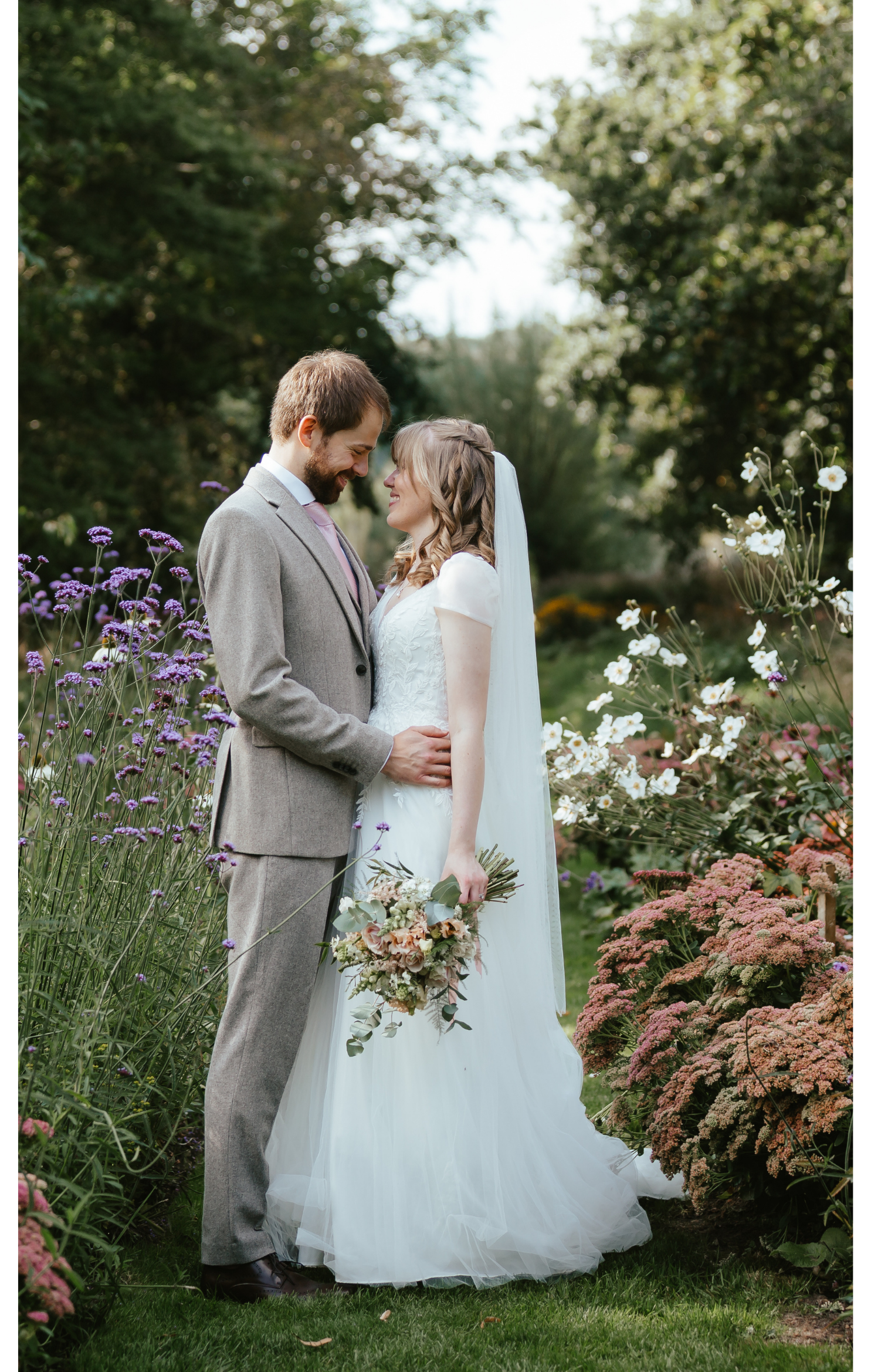 A bride and groom stand close together outdoors in a garden surrounded by blooming flowers. The groom, wearing a light brown suit, is gently holding the bride's waist. The bride, in a white wedding dress, is holding a bouquet of flowers and gazing in