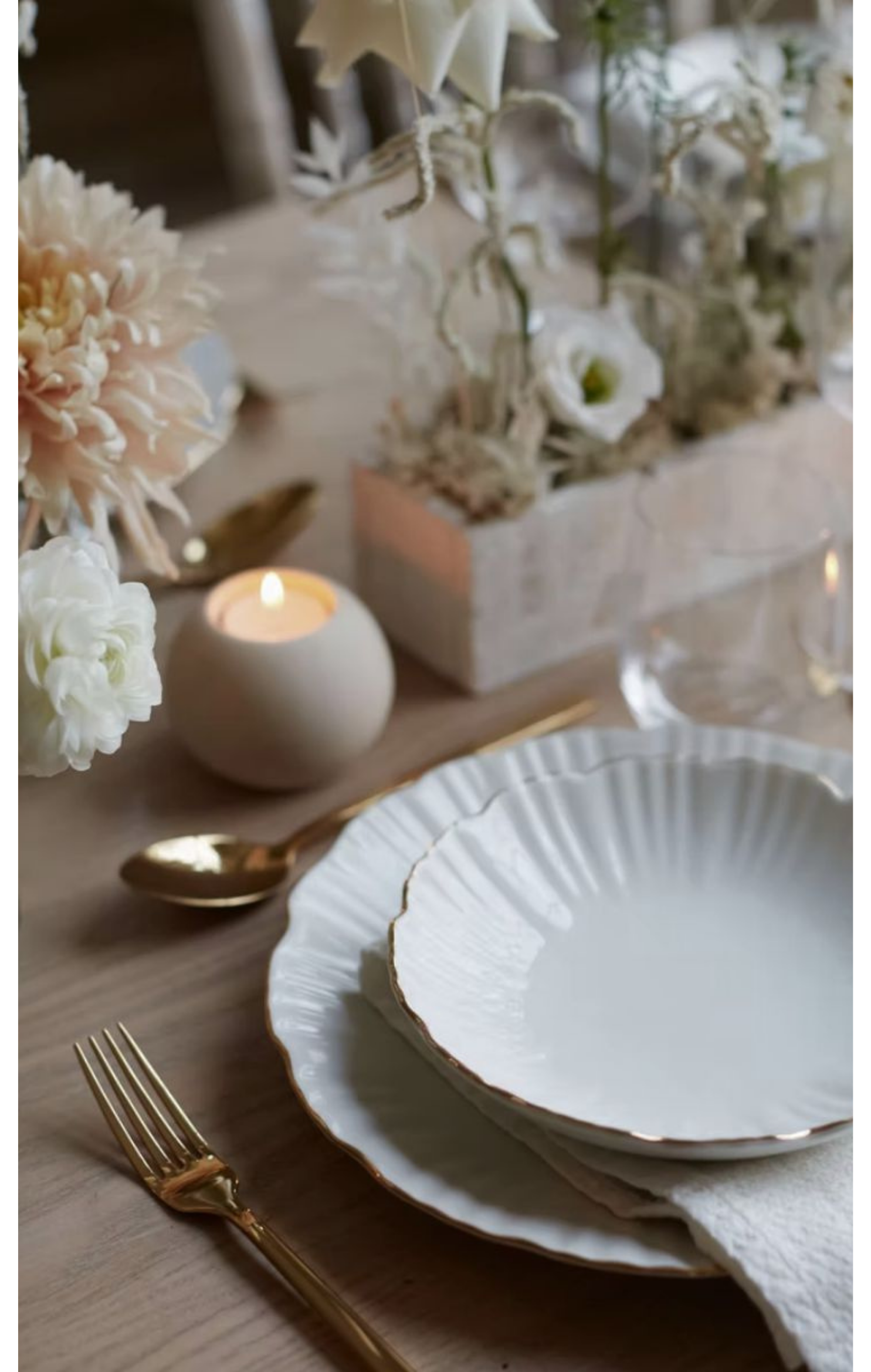 Elegant table setting with white and gold plates, gold fork and spoon, floral centerpiece with white flowers, candle, and soft lighting.