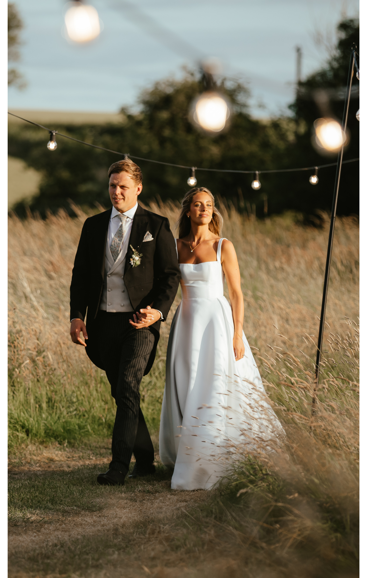A bride and groom walking outdoors in a grassy field, holding hands, with string lights hanging above them during sunset.