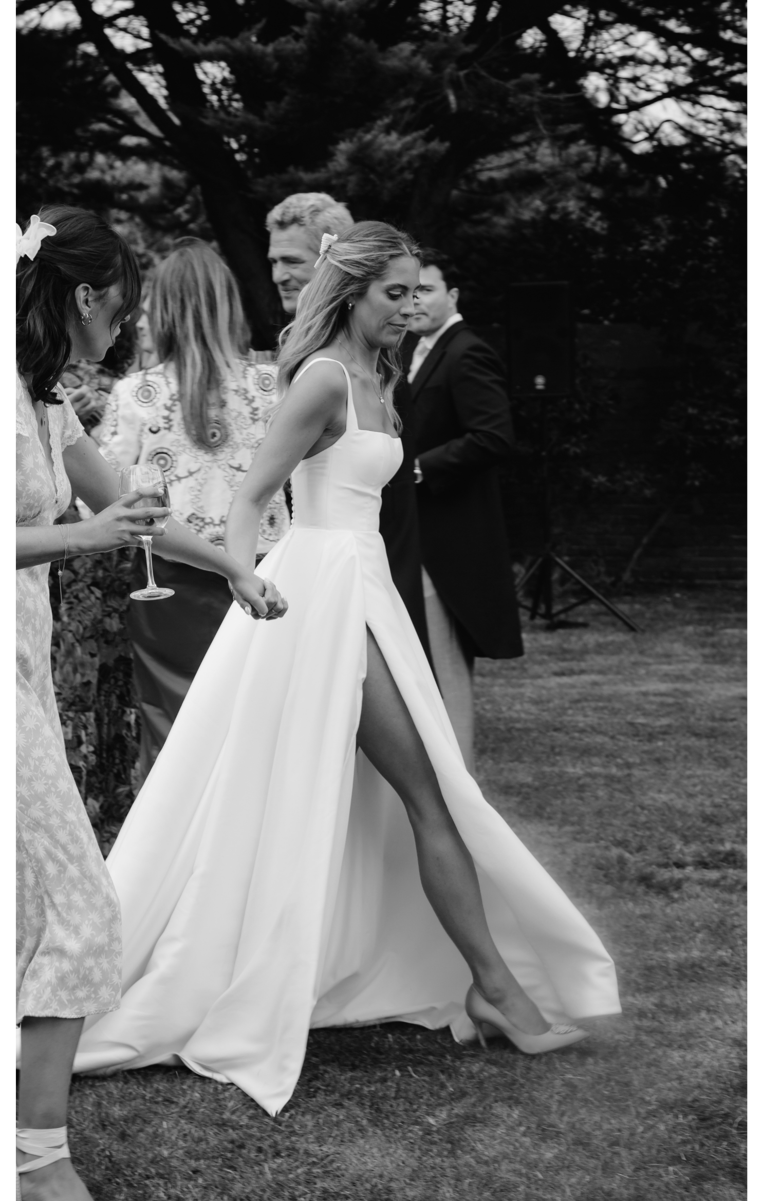 A woman in a long white dress and high heels walking outdoors at a celebration, surrounded by other people, with trees in the background, holding hands with another woman in a patterned dress, one person holding a glass of wine.