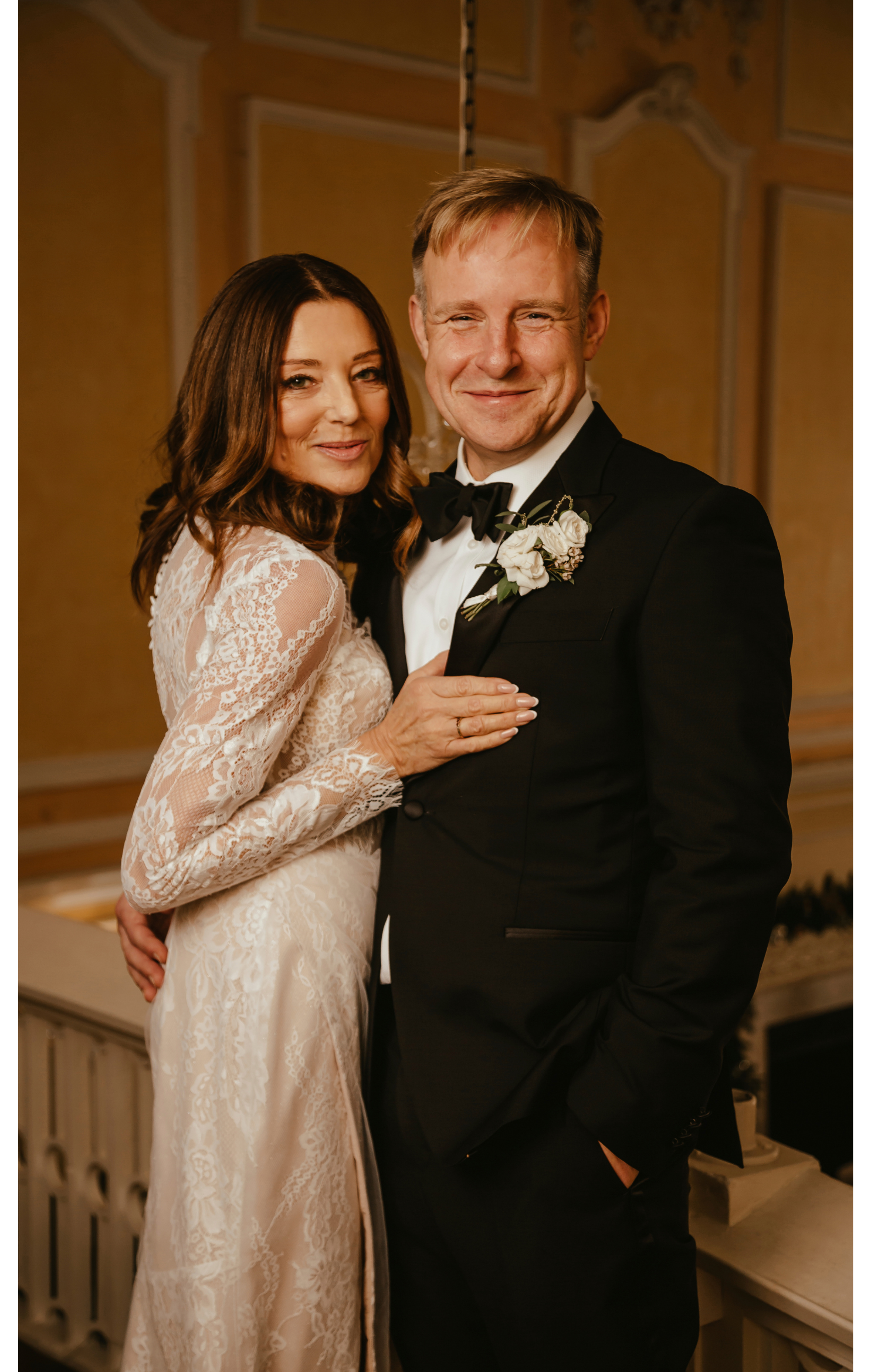 A bride and groom smiling and embracing at their wedding reception, with the bride wearing a lace wedding gown and the groom in a black tuxedo with a bow tie and boutonniere.