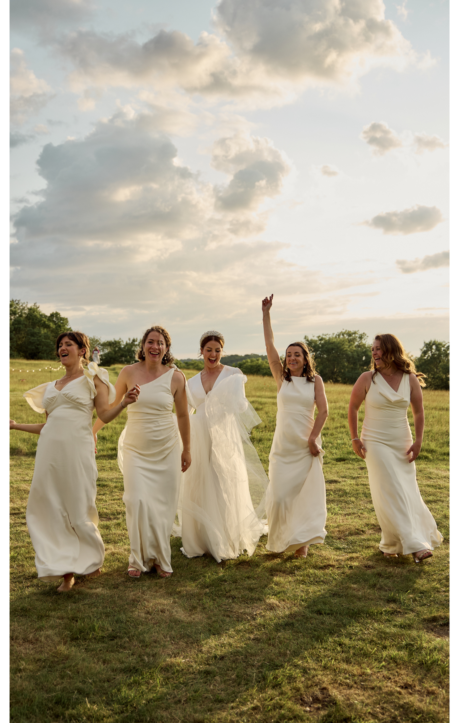 A group of five women in white dresses, possibly bridesmaids, walking outdoors on a grassy field with a cloudy sky in the background during sunset, all smiling and enjoying themselves.