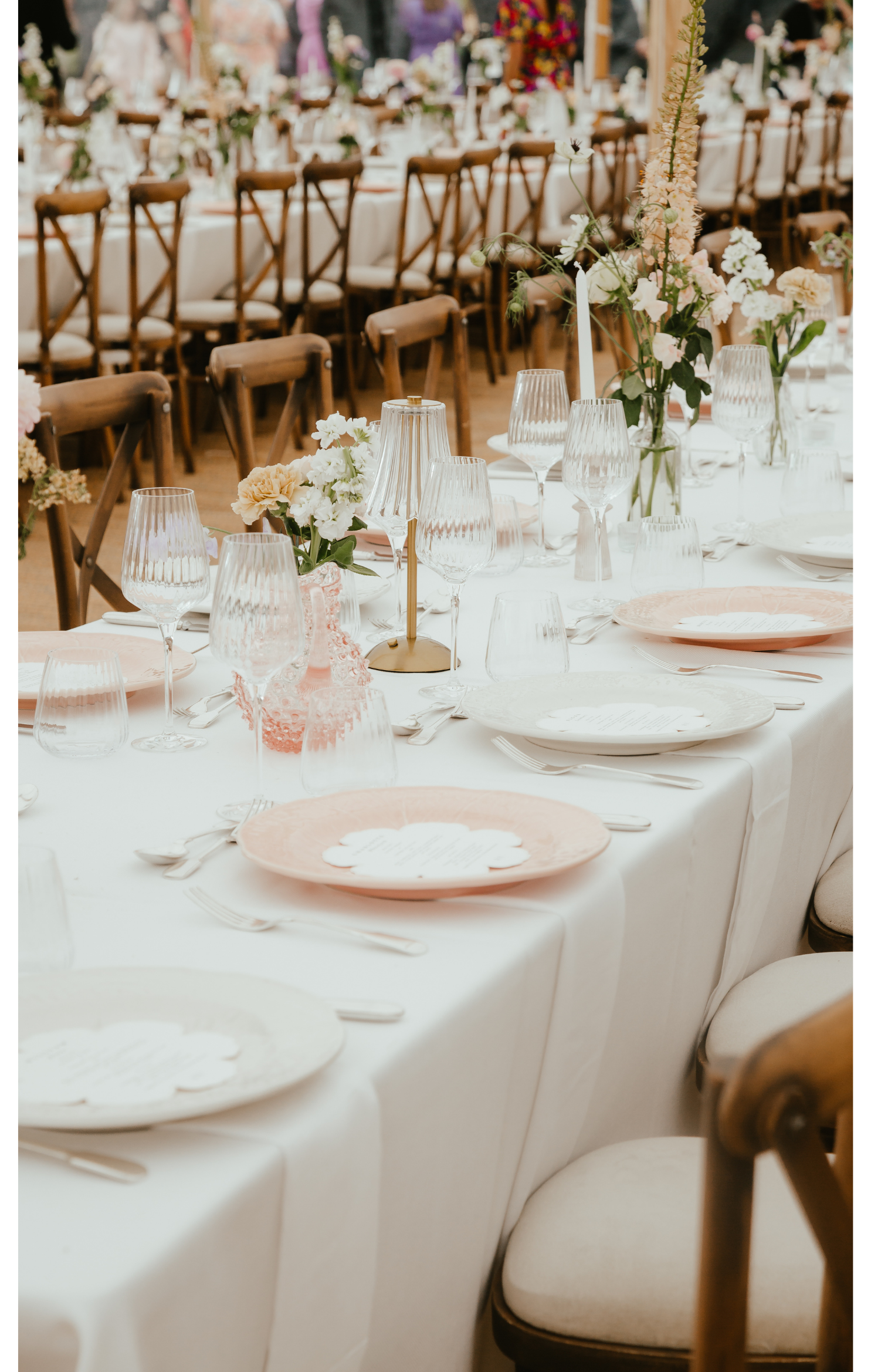 Elegant banquet table set for a wedding or special event with white tablecloths, pink and white floral centerpieces, glassware, and plates, surrounded by wooden chairs.