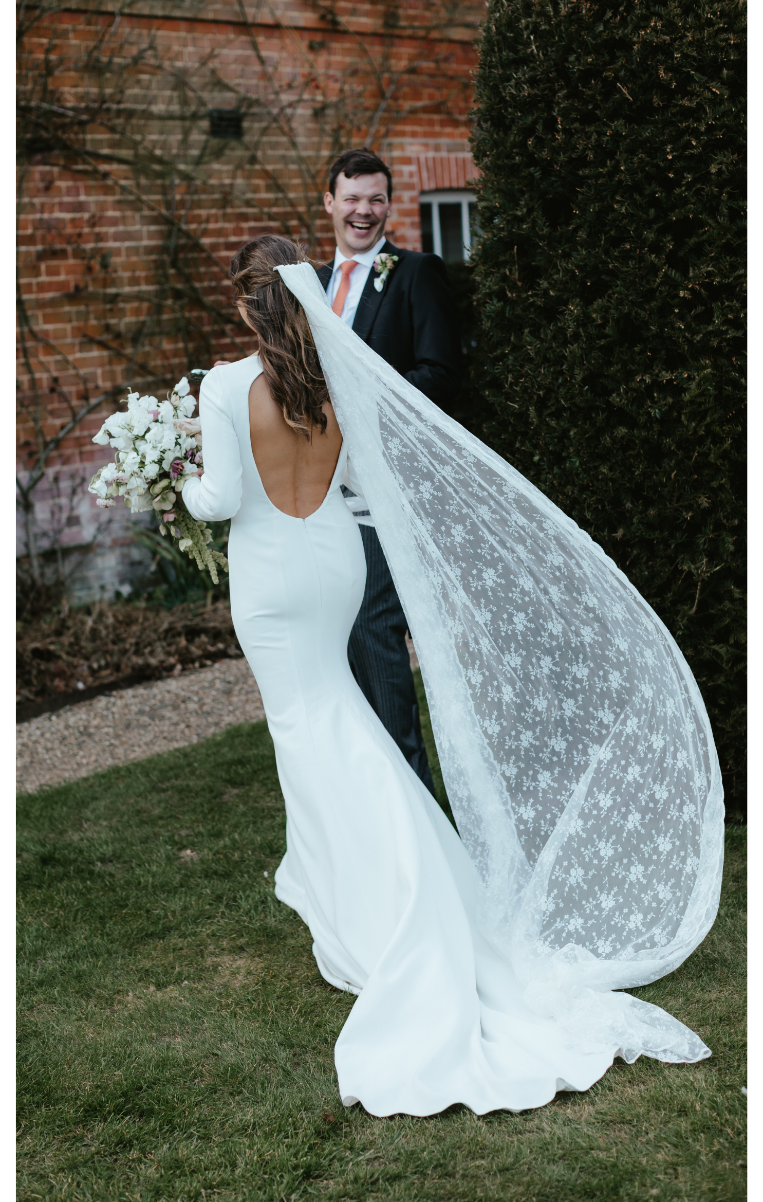 Bride and groom at their wedding outdoors, the bride in a white gown with an open back and floral veil, holding a bouquet, and the groom in a black suit with a boutonniere, smiling.