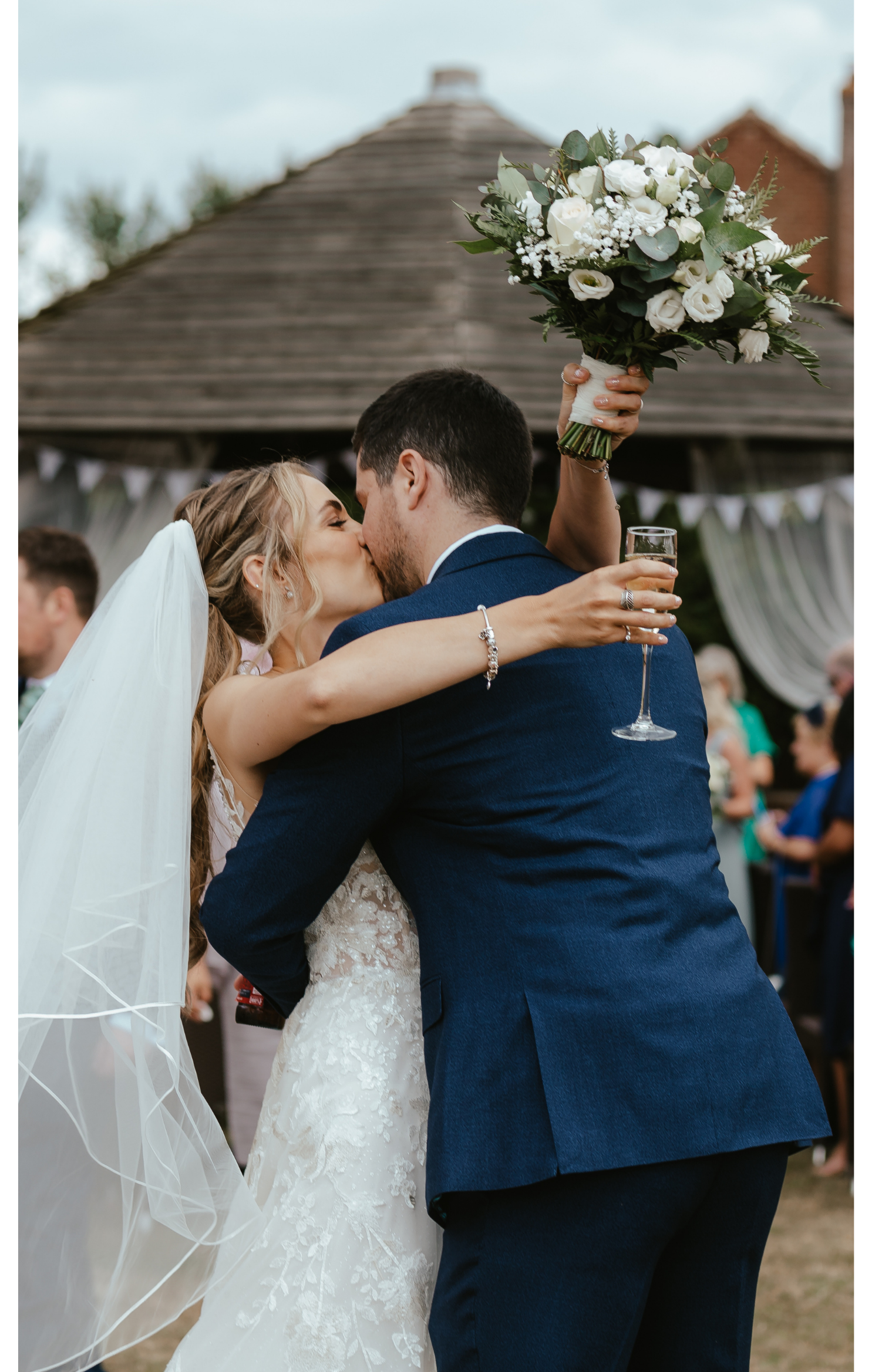 A bride and groom kissing during their wedding celebration, with the bride holding a bouquet of white flowers and the groom holding a glass of champagne.