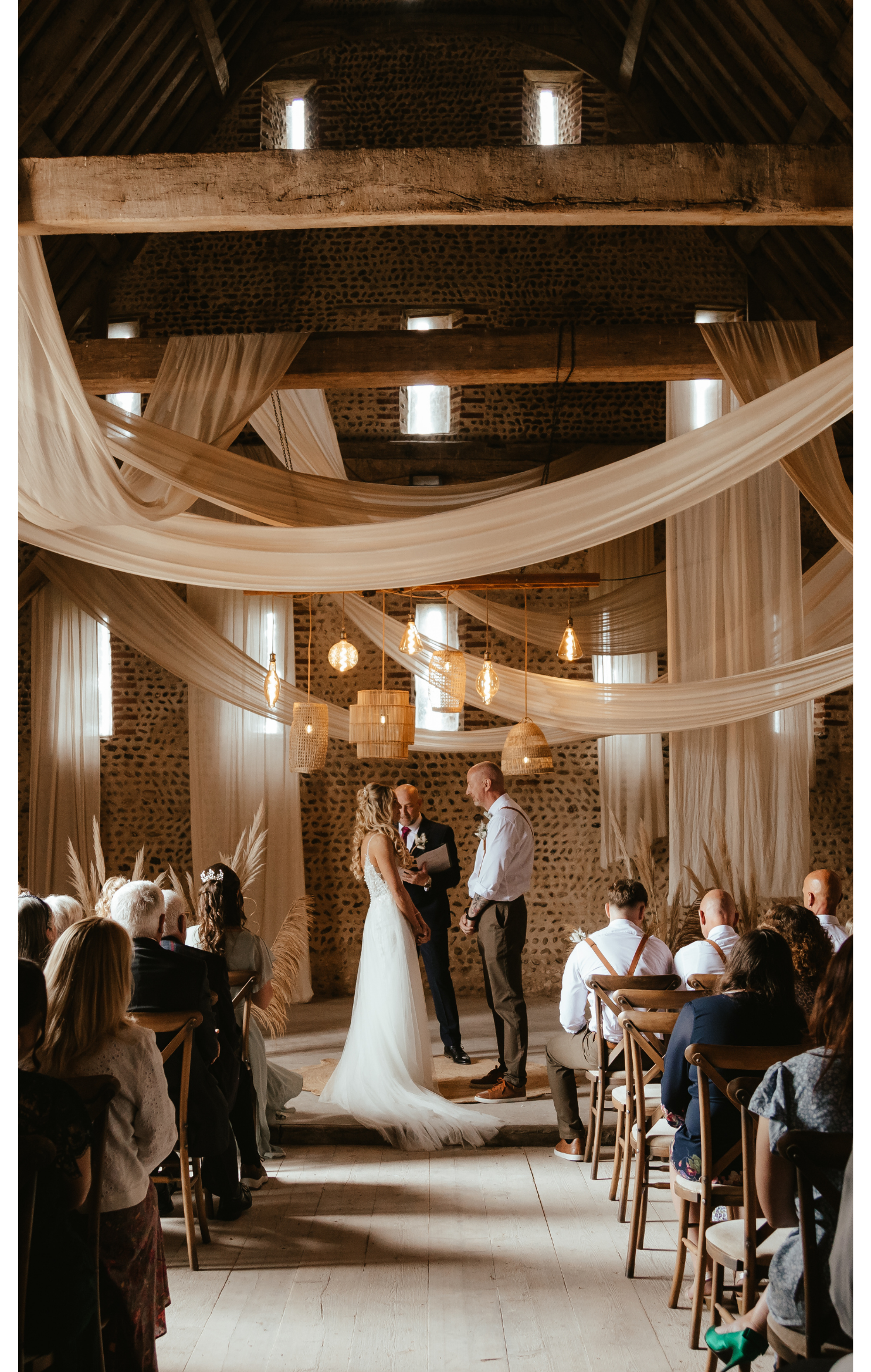 A wedding ceremony in a rustic venue with draped fabric and hanging lights, featuring a bride and groom exchanging vows before an officiant, with seated guests watching.