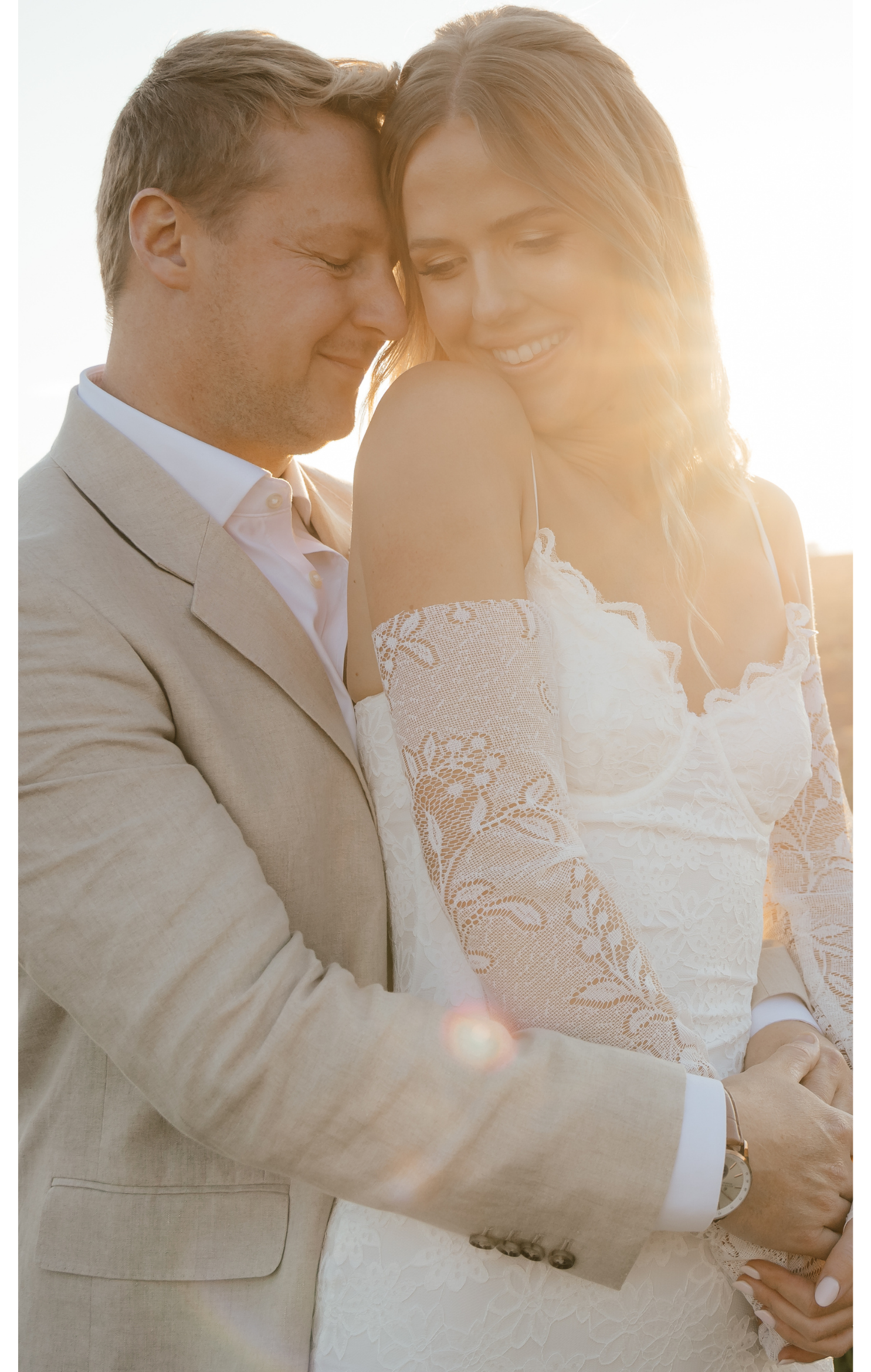 A happy couple embracing outdoors during sunset, with the man in a light beige suit and the woman in a white lace dress with off-the-shoulder sleeves.