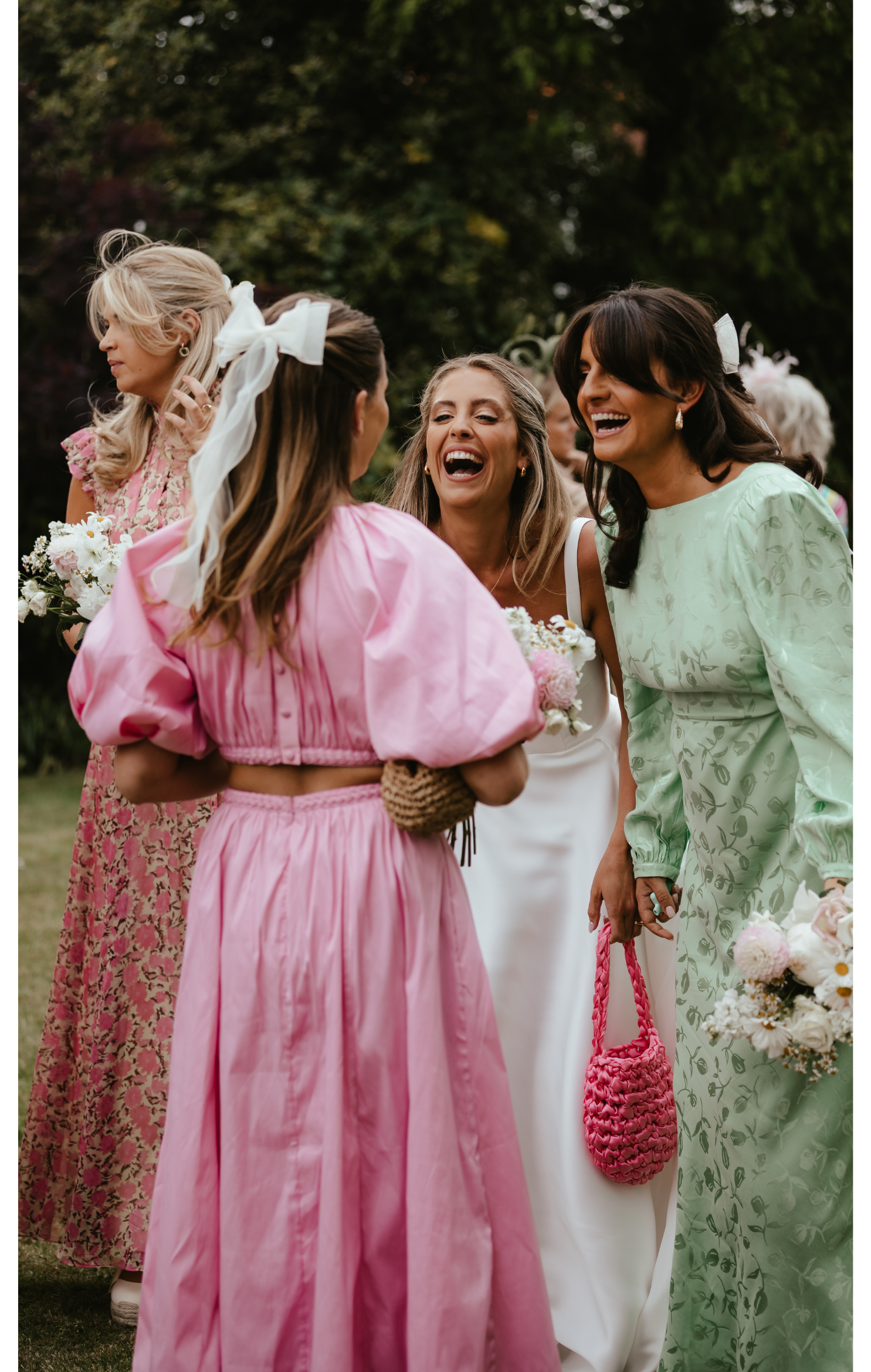 Group of women smiling and talking outdoors during a wedding or event, with some holding flowers and pink woven bags, surrounded by trees and greenery.