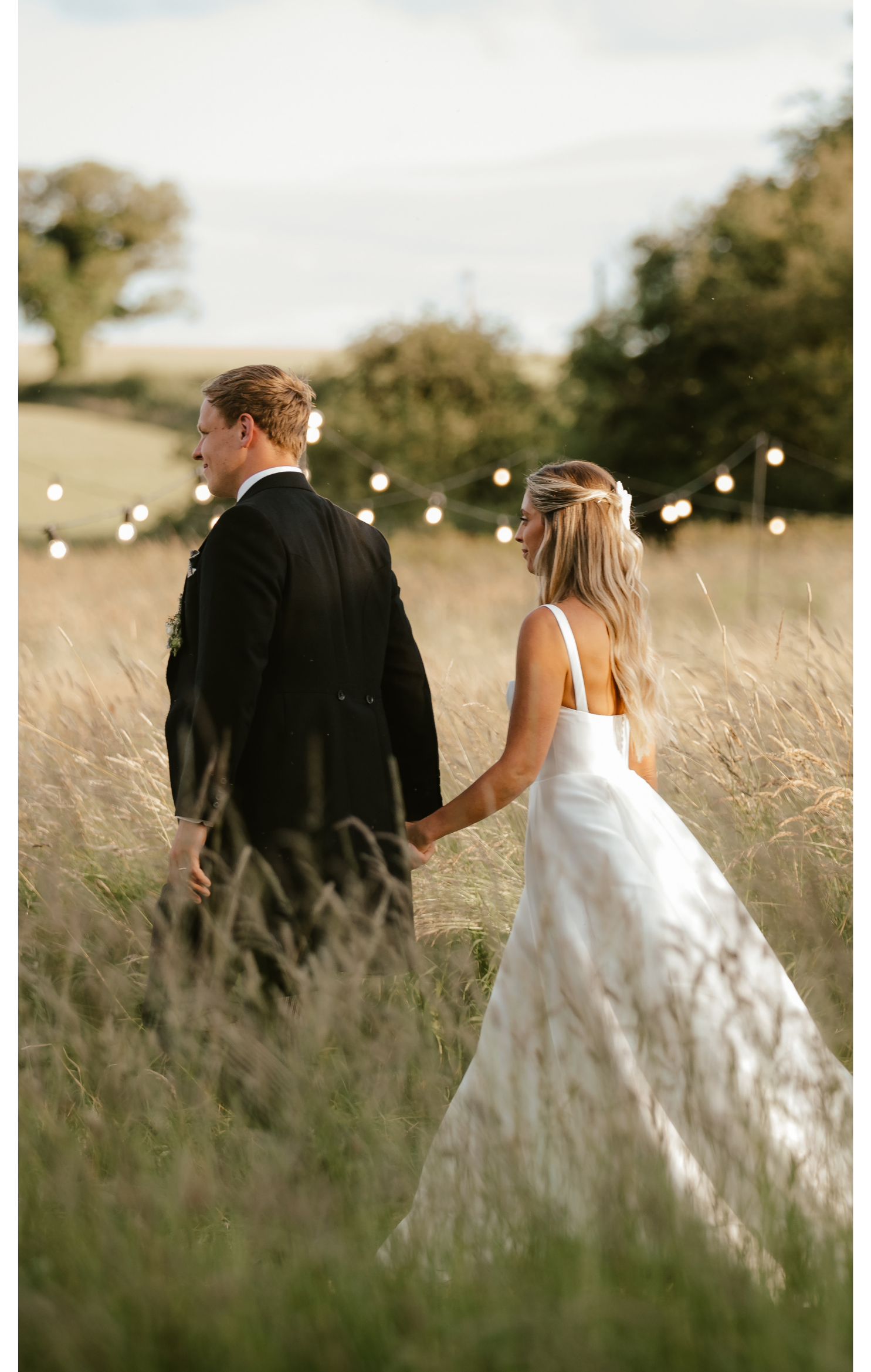 A bride and groom holding hands in a grassy field during sunset, with string lights hanging behind them.